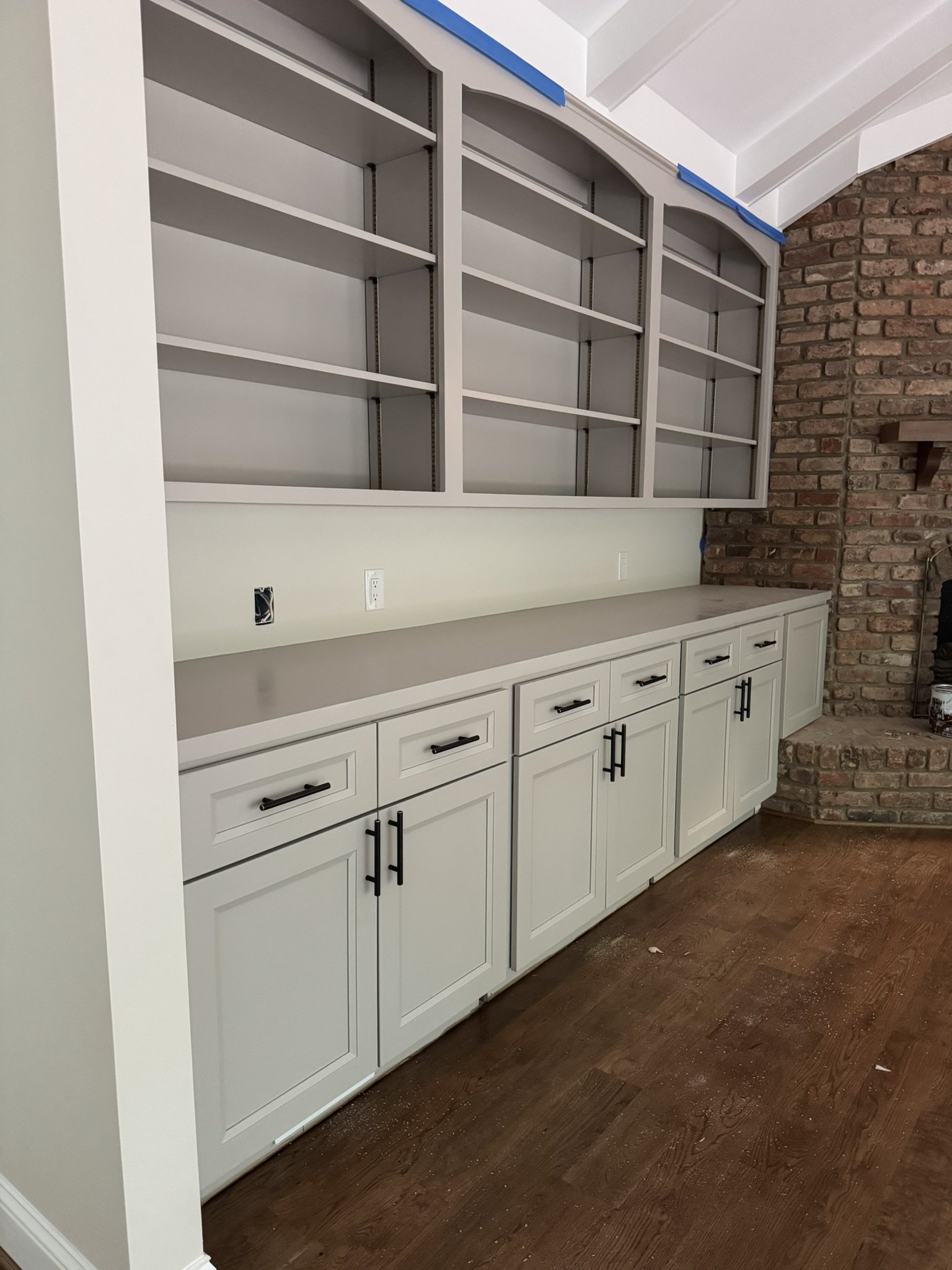 White built-in kitchen cabinet with glass doors, black handles, and drawers, under a sloped ceiling with brick wall on the side, and a brown wooden floor.