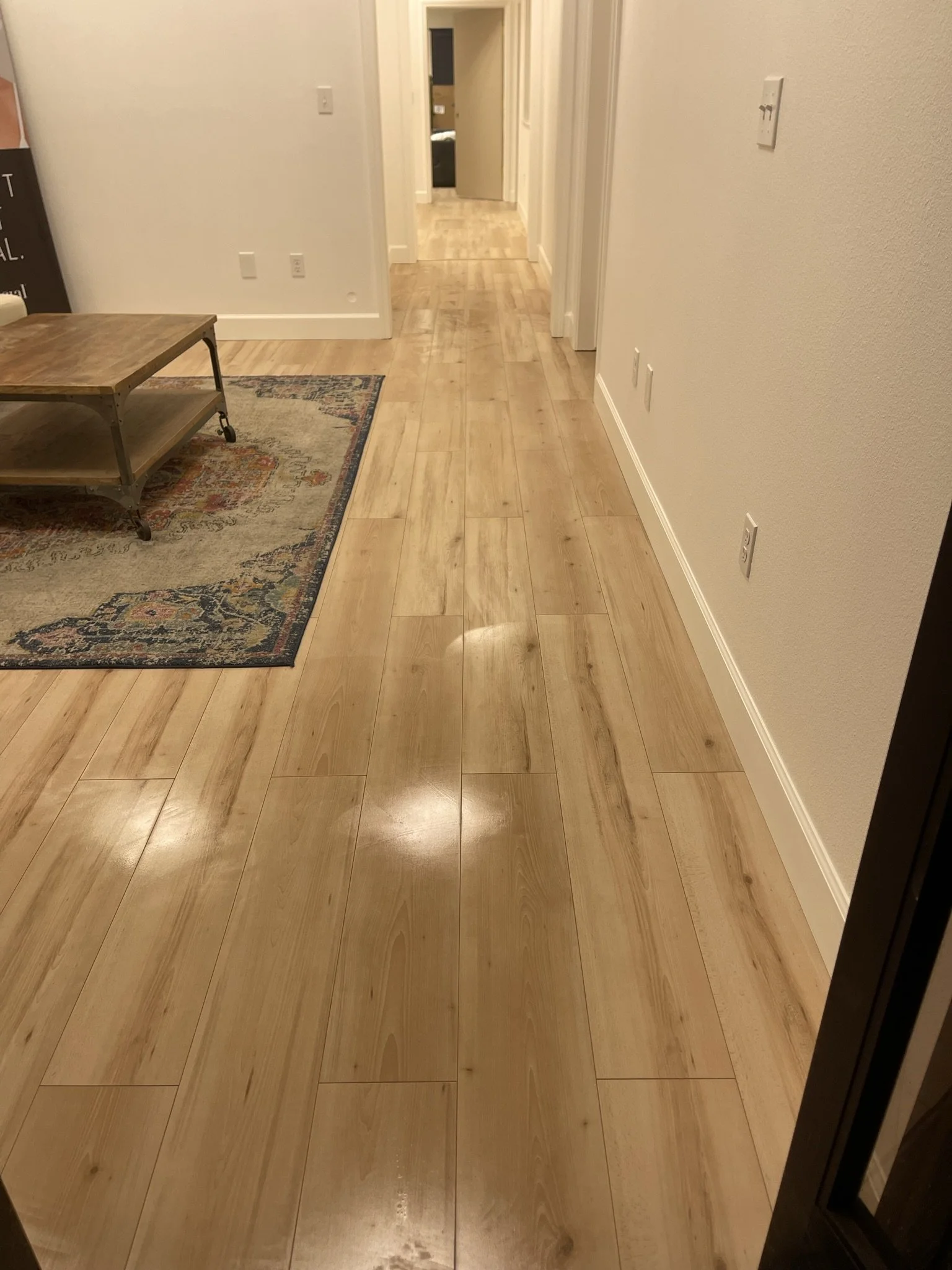 Wooden floor hallway leading to a door in the distance with a picture on the left side. There are multiple wall outlets and switches along the walls, and a small wooden table with wheels on a patterned rug on the left side.
