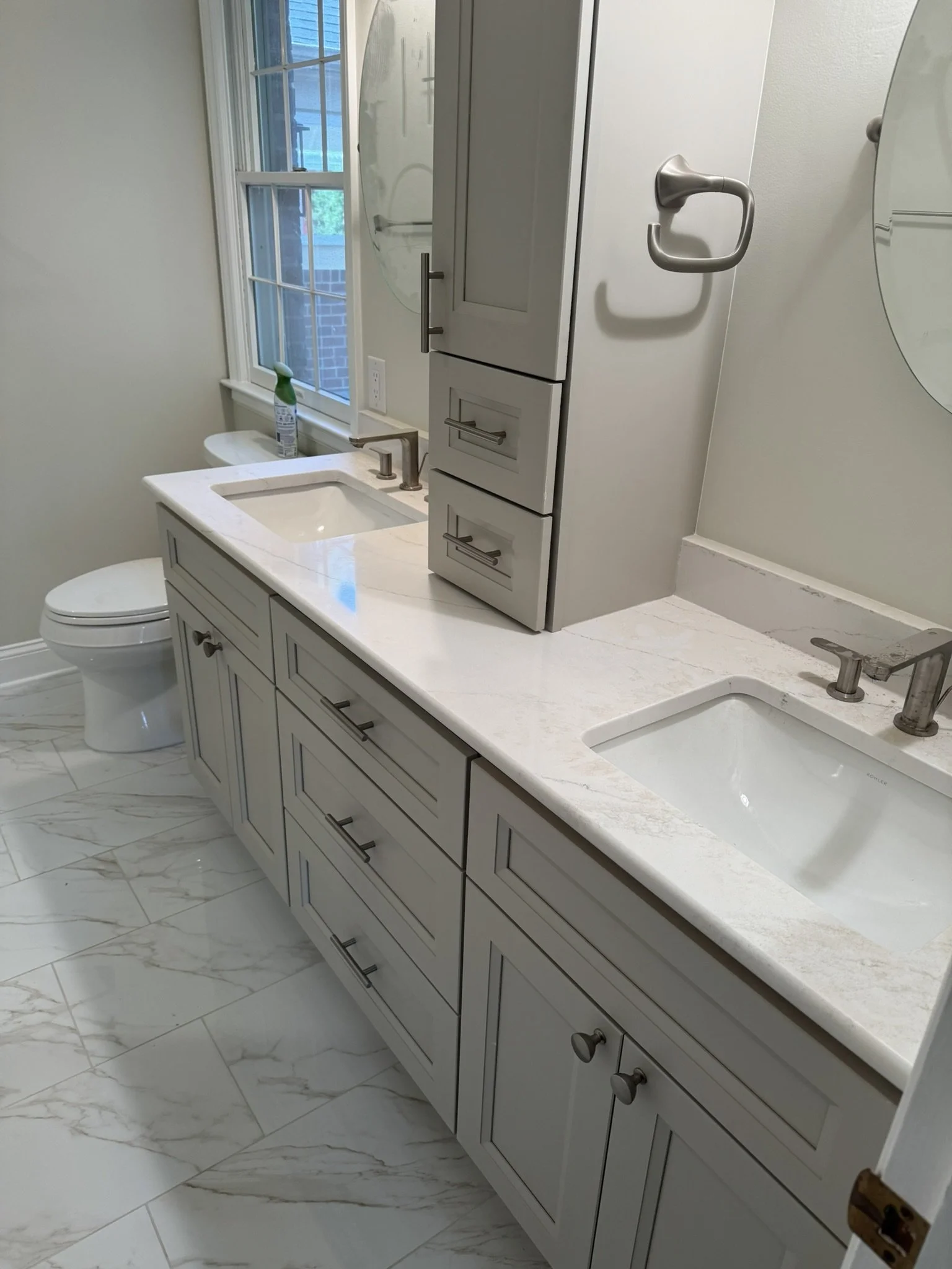 A modern bathroom with white cabinets, marble countertop, and a large window with a view of trees outside.
