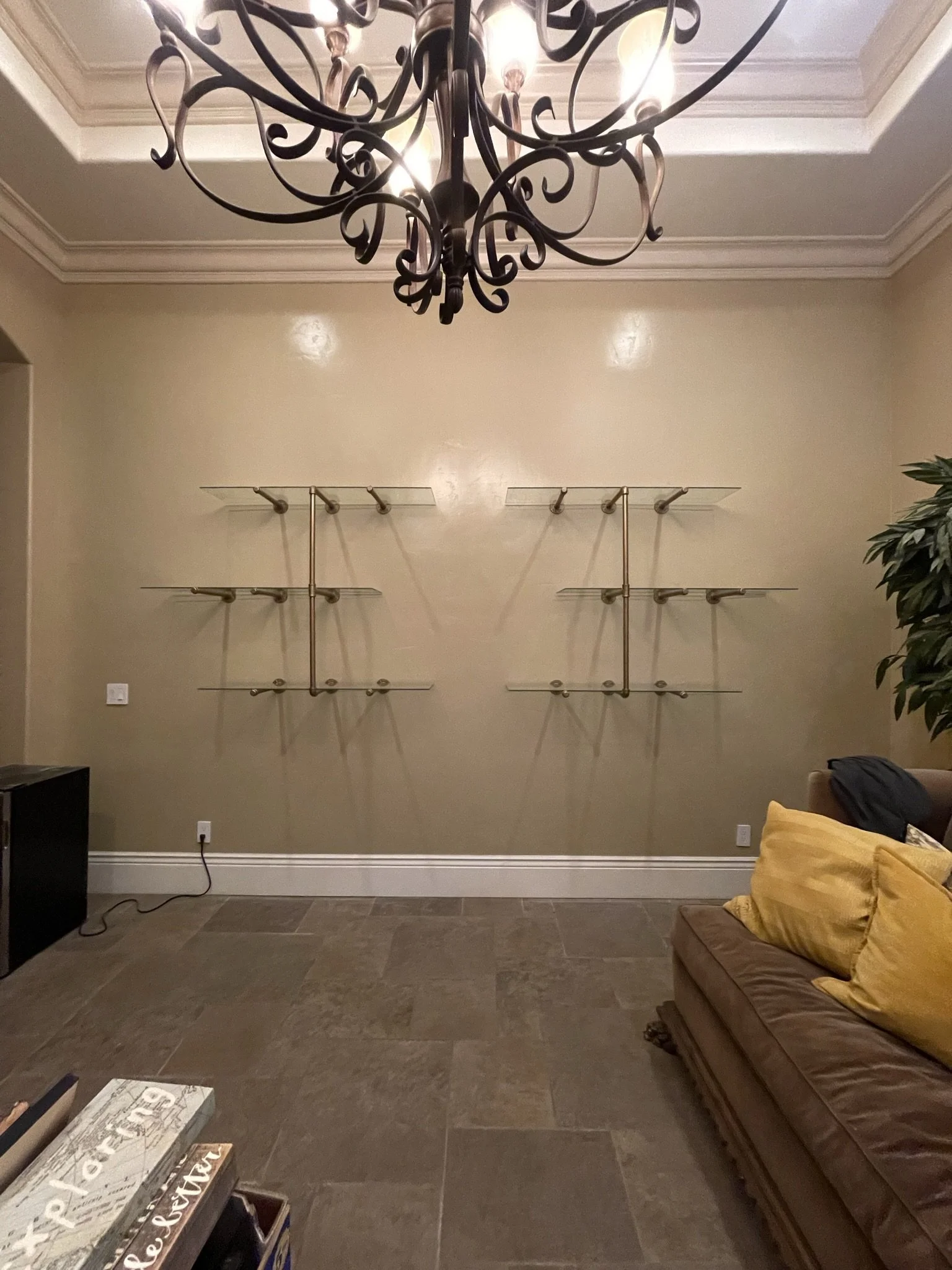 Living room with beige walls, a dark brown couch with yellow pillows, a chandelier hanging from the ceiling, a decorative shelving unit on the wall, and a plant in the corner.