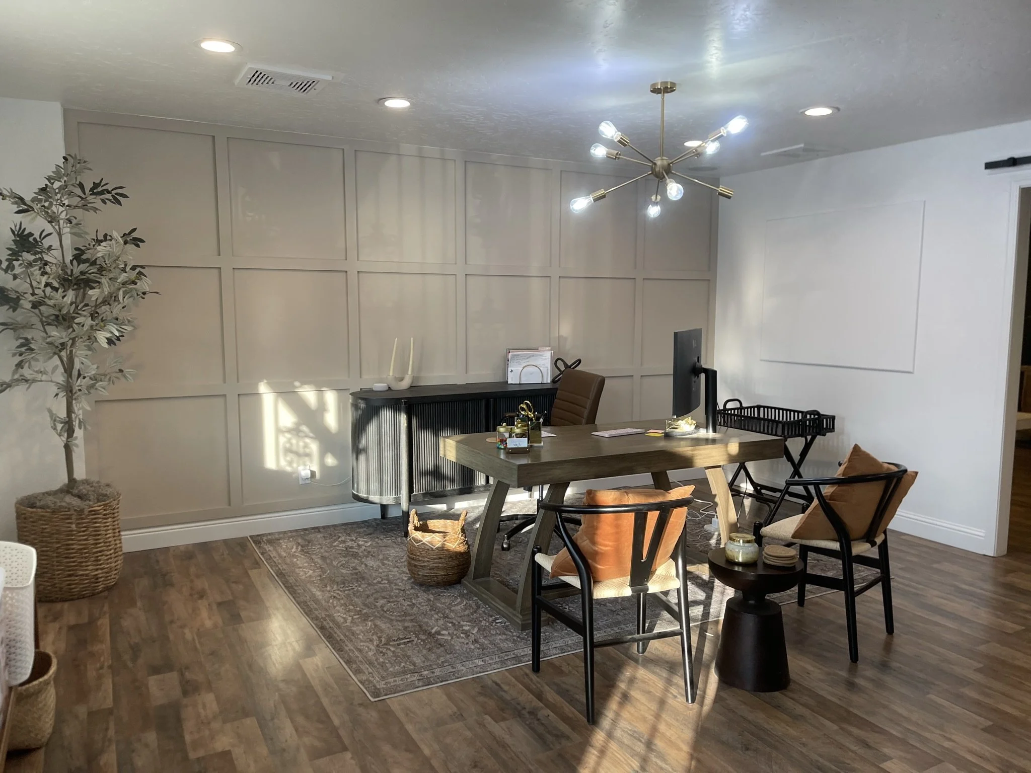 Modern office with white paneled wall, wooden desk, black office chair, plants, decorative items, and ceiling light fixtures.