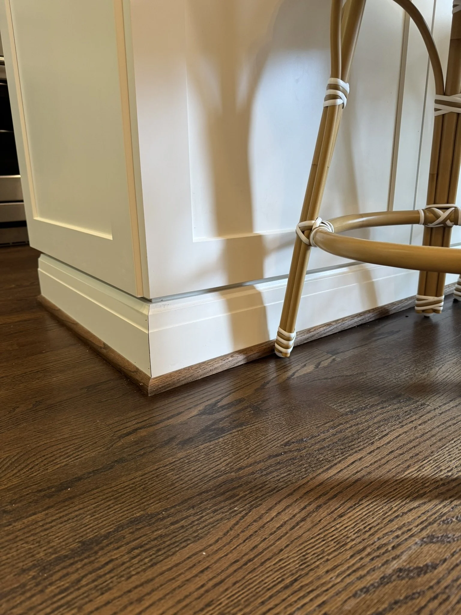 Close-up of a corner of a white kitchen cabinet with a wooden floor, and part of a rattan chair with white ties.