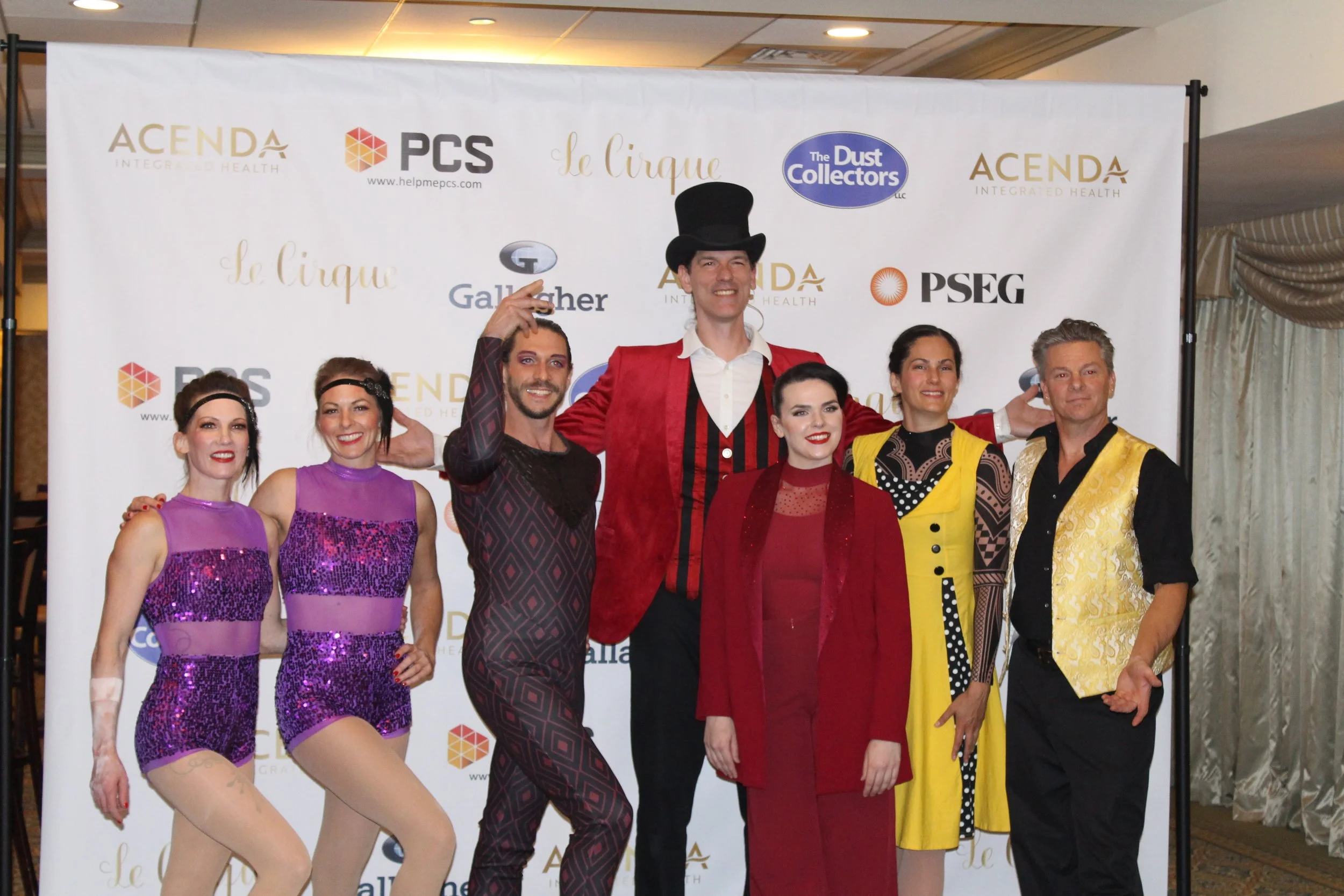 Le Cirque performers pose in front of step and repeat backdrop featuring sponsor logos 