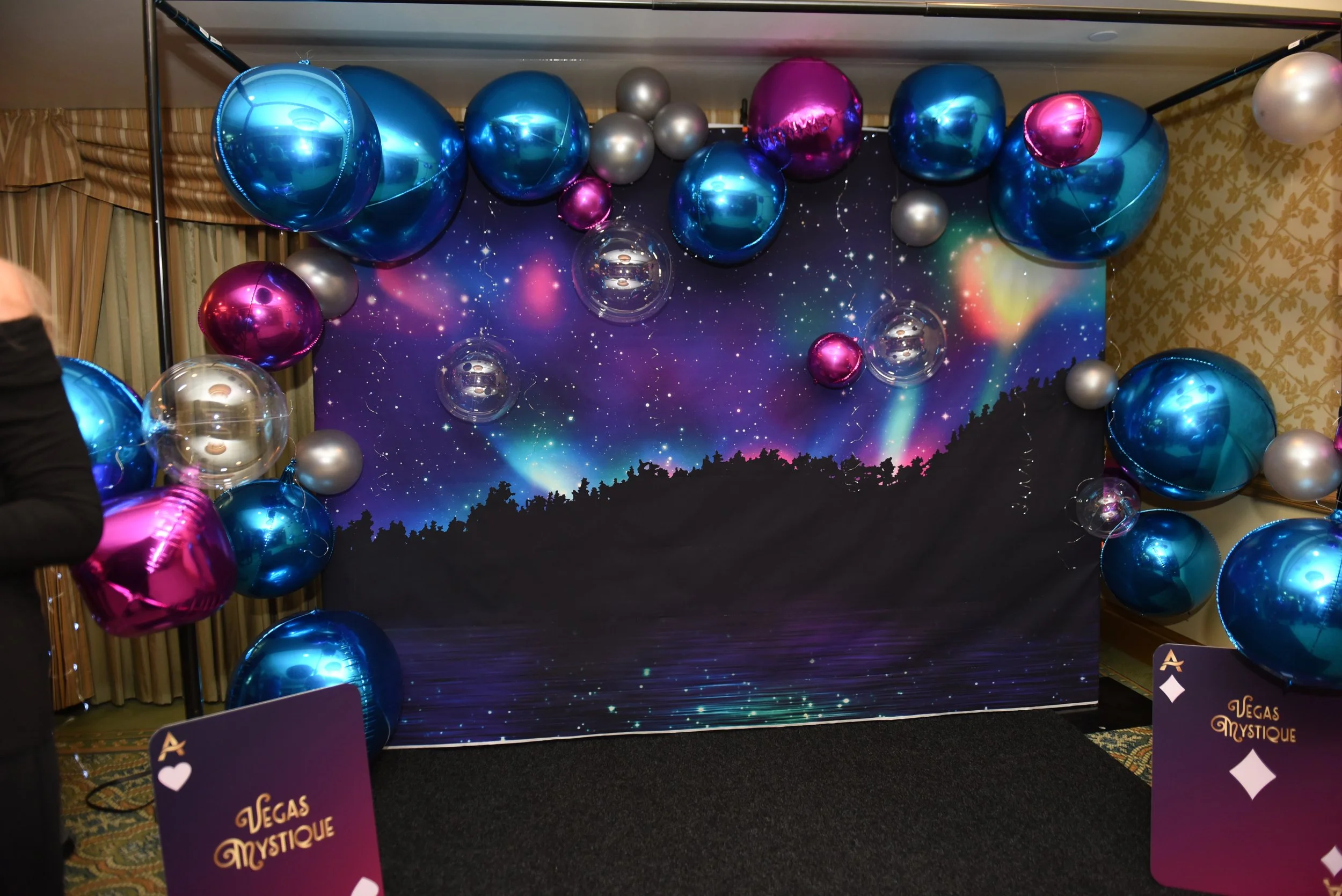 Photo backdrop featuring a nighttime image of a lake forest with northern light streams in pink and blue with pink, blue, and sparkly balloon decor and vegas mystique cards