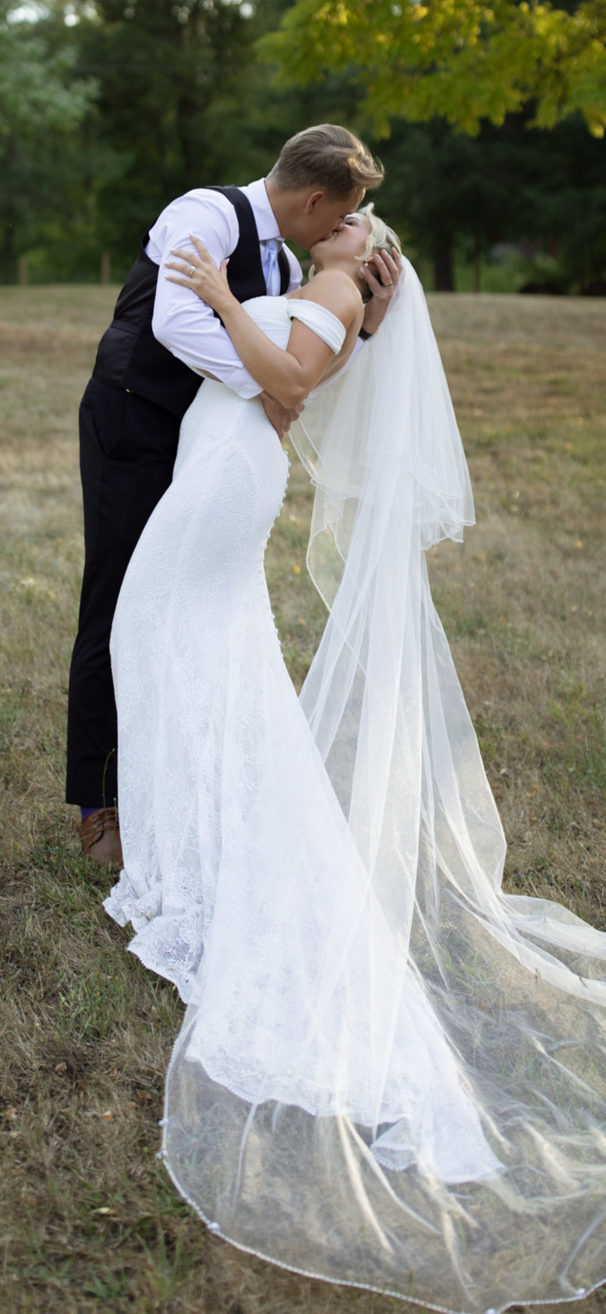 A bride and groom kissing outdoors during their wedding. The bride is wearing a white wedding gown with a long veil, and the groom is dressed in a white shirt with a black vest and pants. They are standing on grass with trees in the background.
