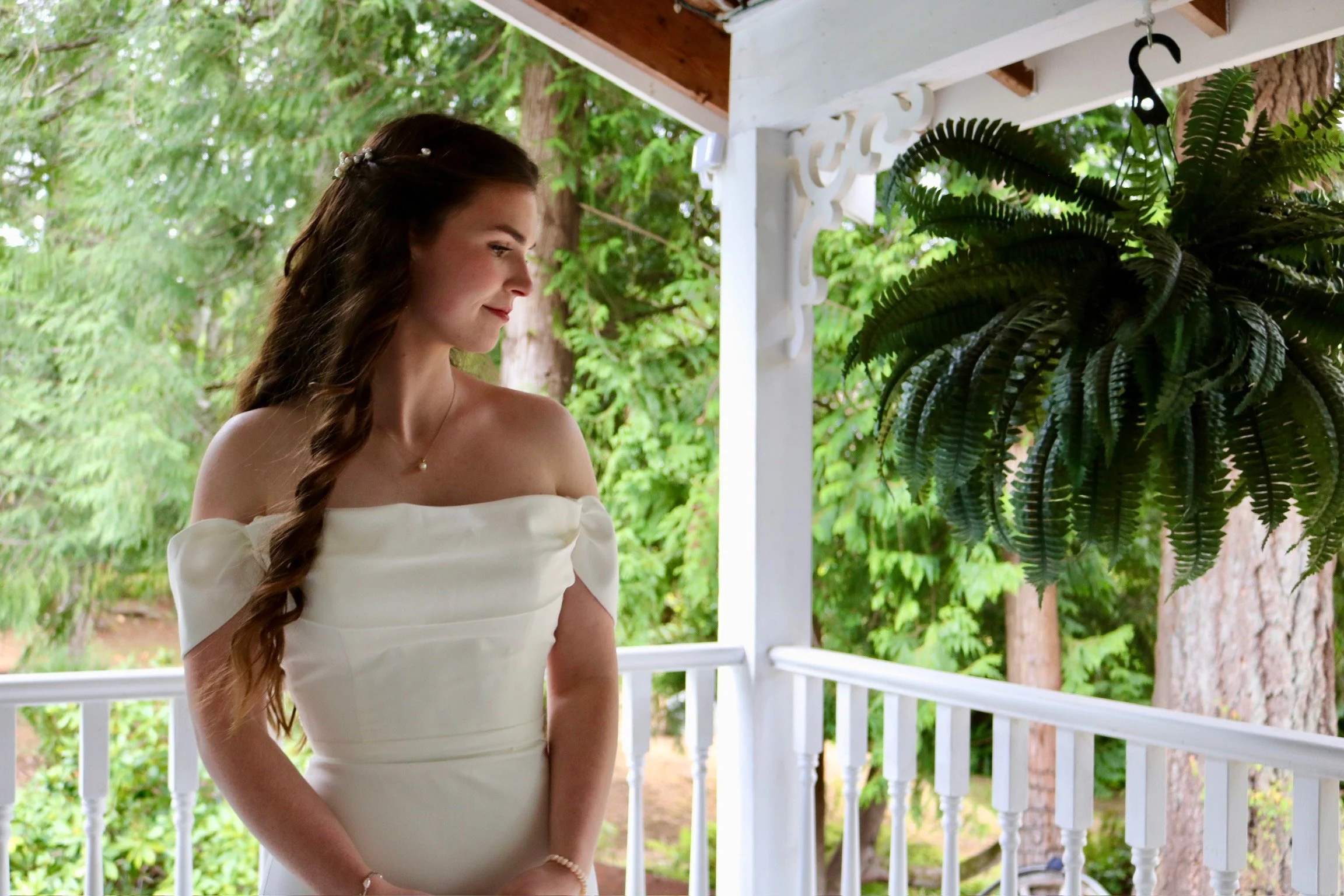 A young woman with long, dark, curled hair and a pearl hair accessory, wearing an off-the-shoulder white dress, stands on a porch with white railings, surrounded by green trees and plants, smiling softly.