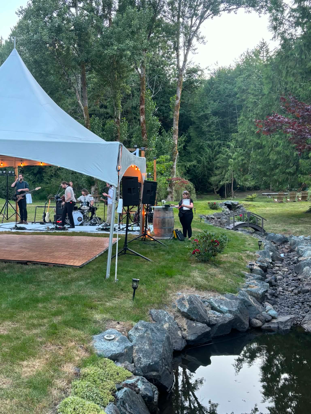 An outdoor band performing on a stage under a white canopy, with a woman standing nearby with musical notes in a park-like setting, surrounded by trees and a small stream with rocks.