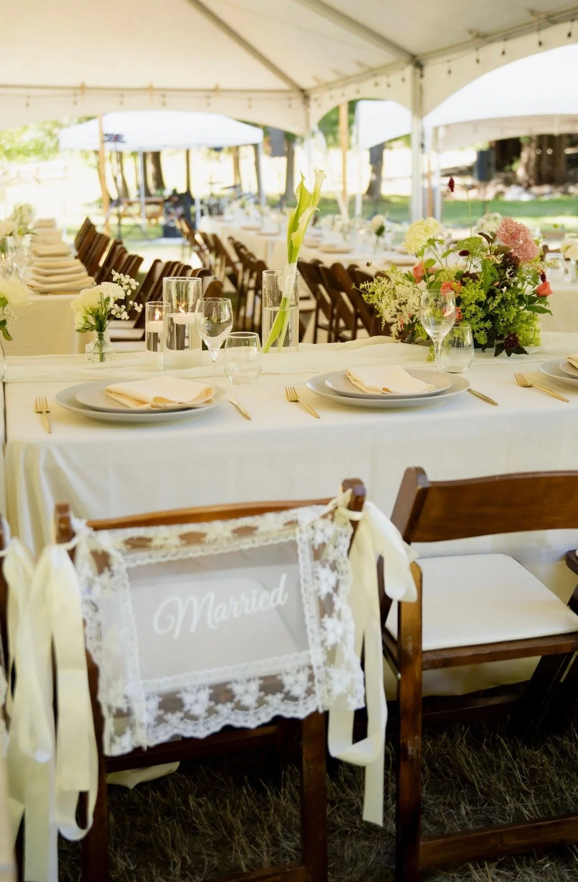 An outdoor wedding reception setup with decorated tables under a tent, featuring floral centerpieces, wine glasses, and place settings, with a 'Married' sign on a chair at the front.