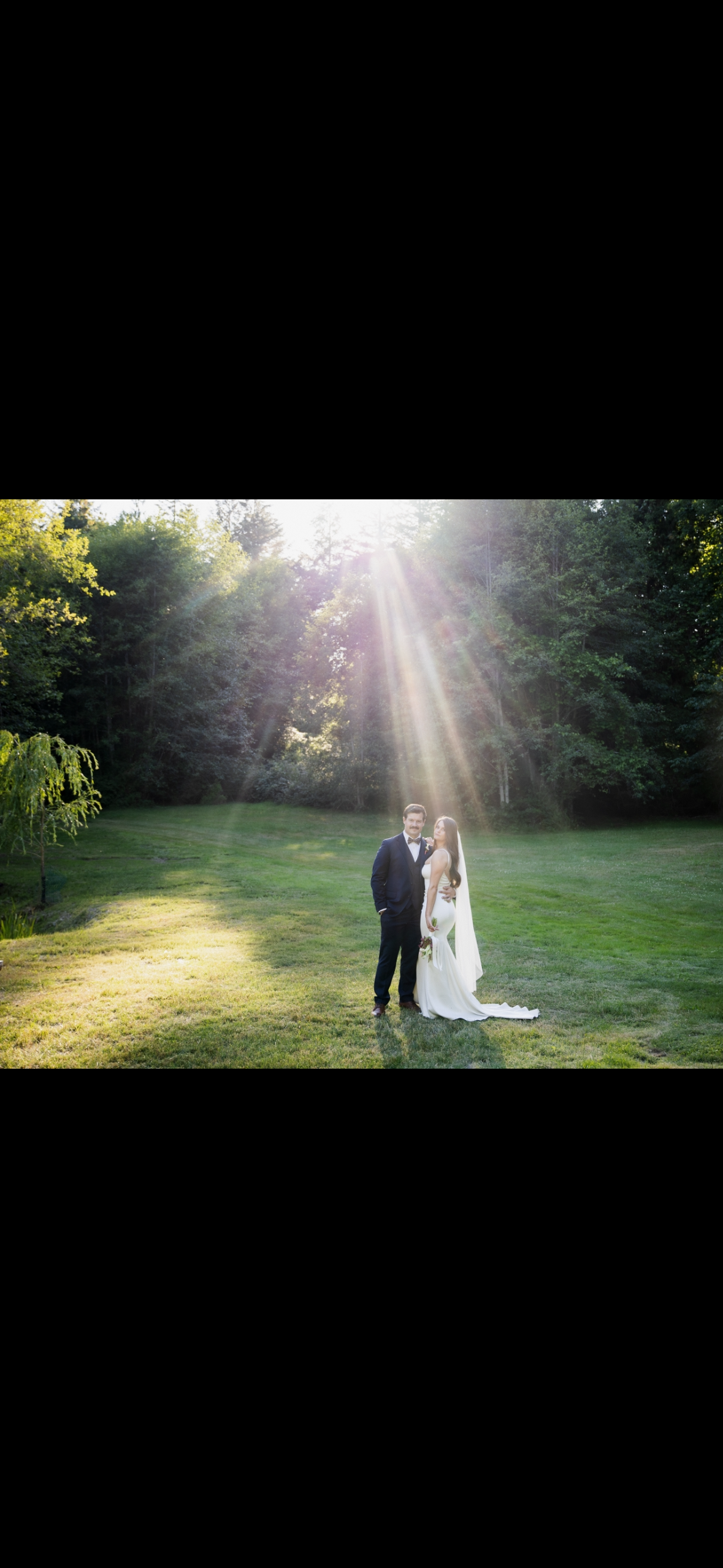 A bride and groom standing on a grassy field with sunlight streaming through the trees overhead, in a scenic outdoor setting.