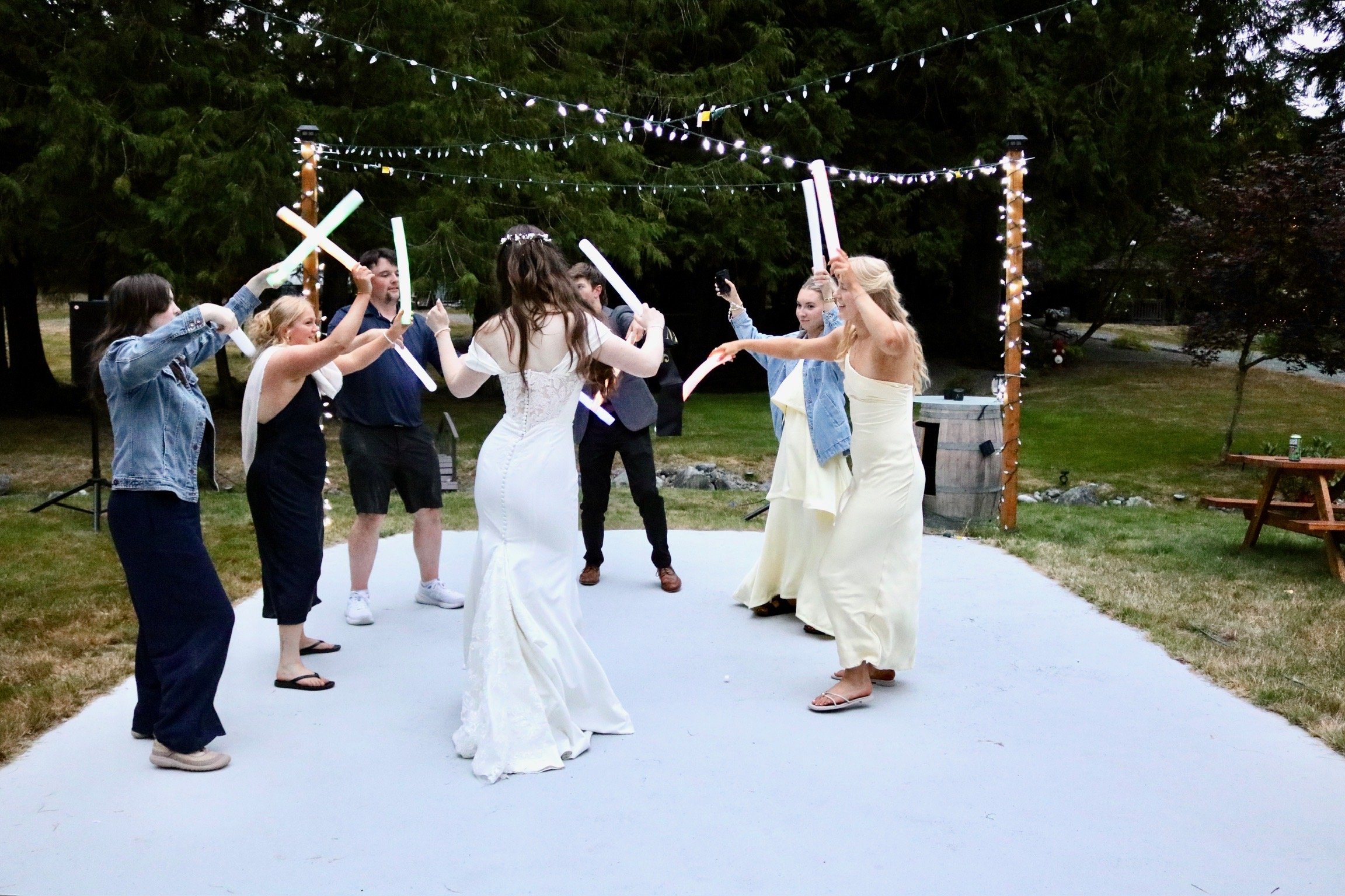 A group of people dancing at an outdoor event, with a woman in a white dress in the center and others holding colorful glow sticks, under string lights in a garden setting during evening.