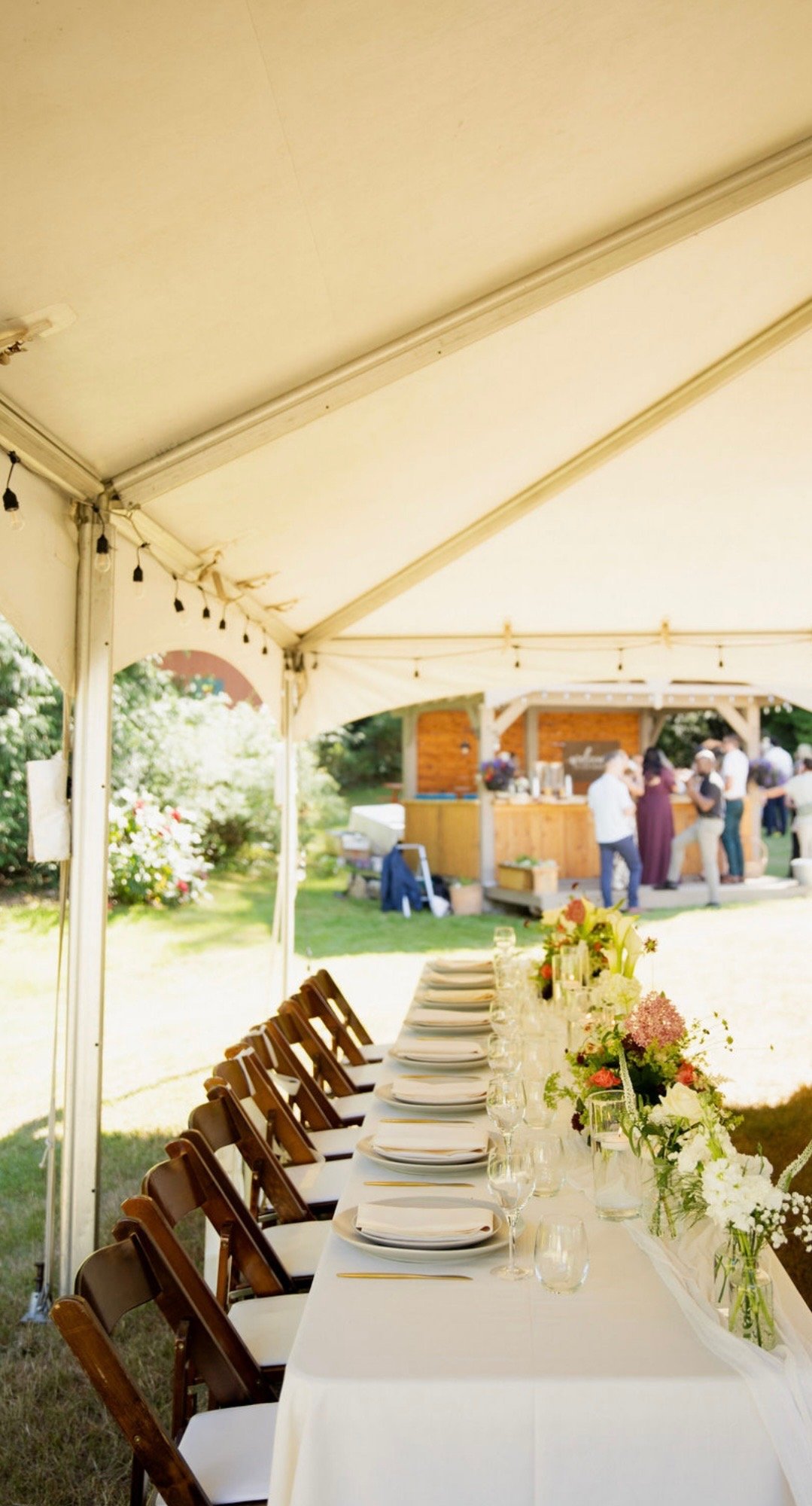 Long outdoor dining table set under a white canopy tent with white plates, glassware, and floral centerpieces, with a garden and a wooden food stand in the background where people are gathered.