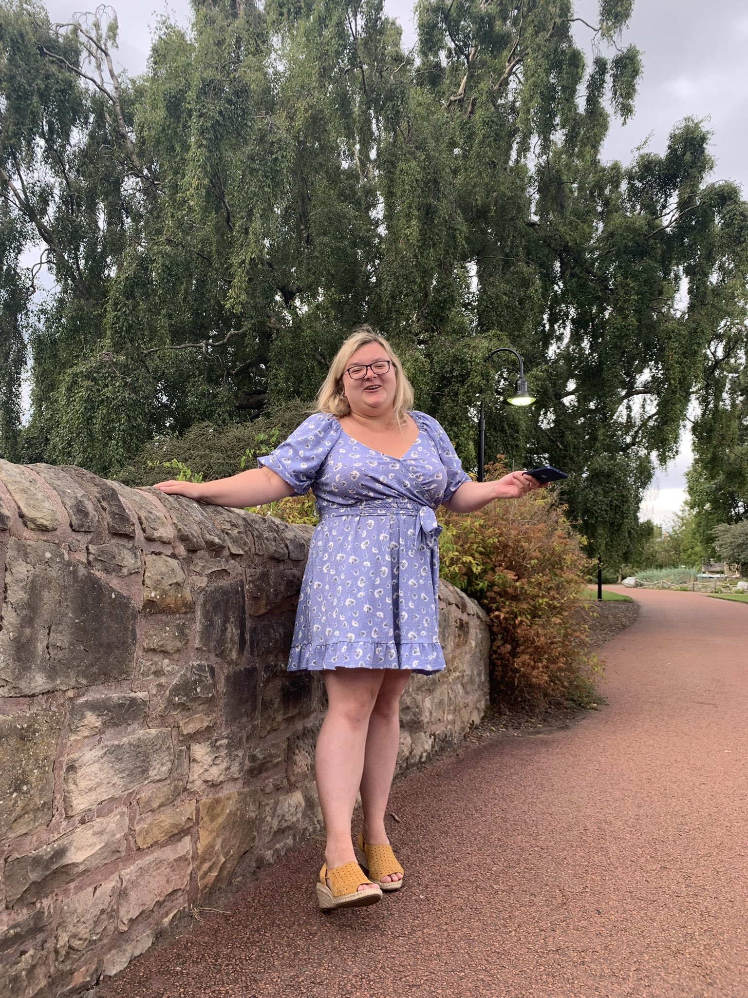 A woman with blonde hair and glasses wearing a blue dress with white patterns, standing outdoors beside a stone wall and holding a phone in her right hand. She is smiling and wearing yellow high-heeled shoes. There are trees and a pathway in the background.