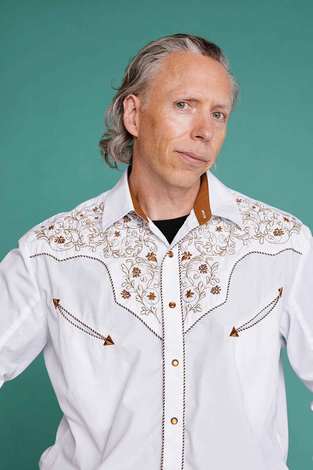 A middle-aged man with gray hair and light skin, wearing a white embroidered western shirt with brown accents, stands against a teal background.