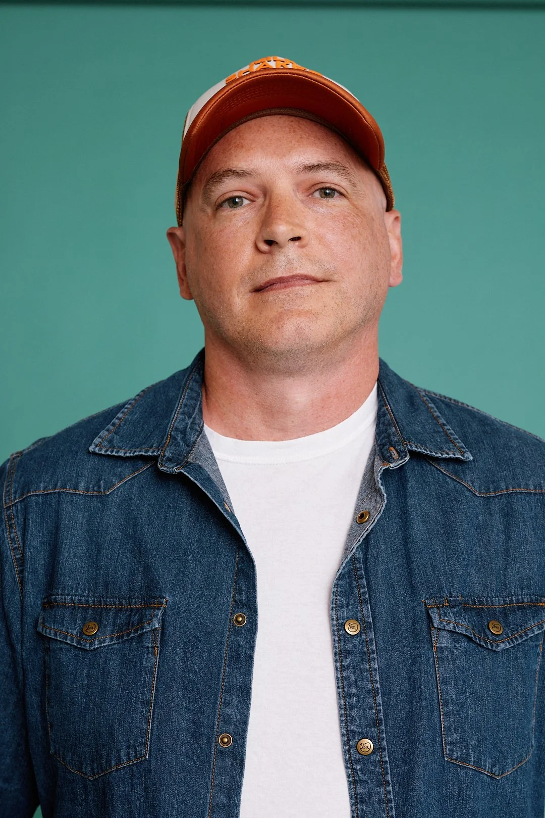 Portrait of a man wearing a denim jacket, white t-shirt, and a brown baseball cap, against a plain teal background.