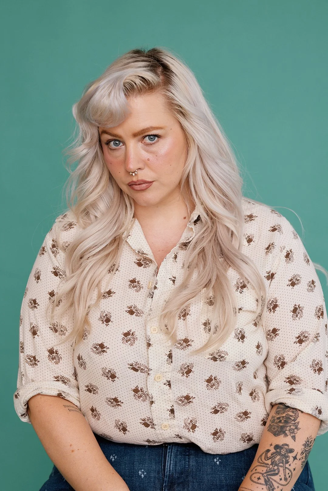 A woman with long, wavy platinum blonde hair and blue eyes, wearing a light-colored floral blouse with rolled-up sleeves, standing against a teal background.
