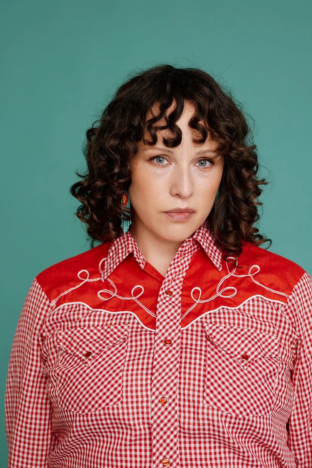 A woman with curly dark hair and blue eyes wearing a red and white checkered western shirt with embroidery and beaded earrings against a teal background.