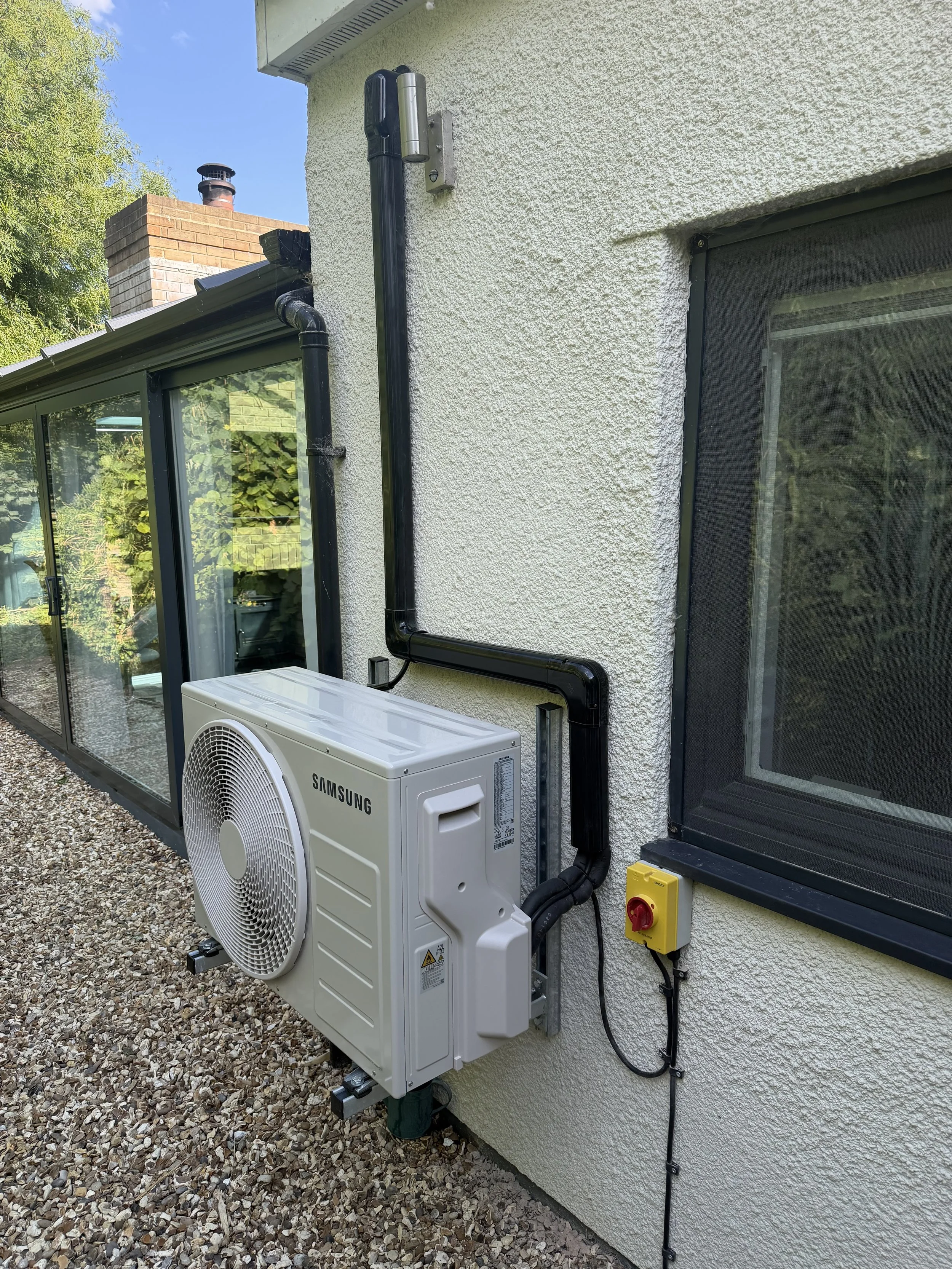 External view of a house showing an air conditioning unit and ductwork on the wall, with a window and gravel ground outside.