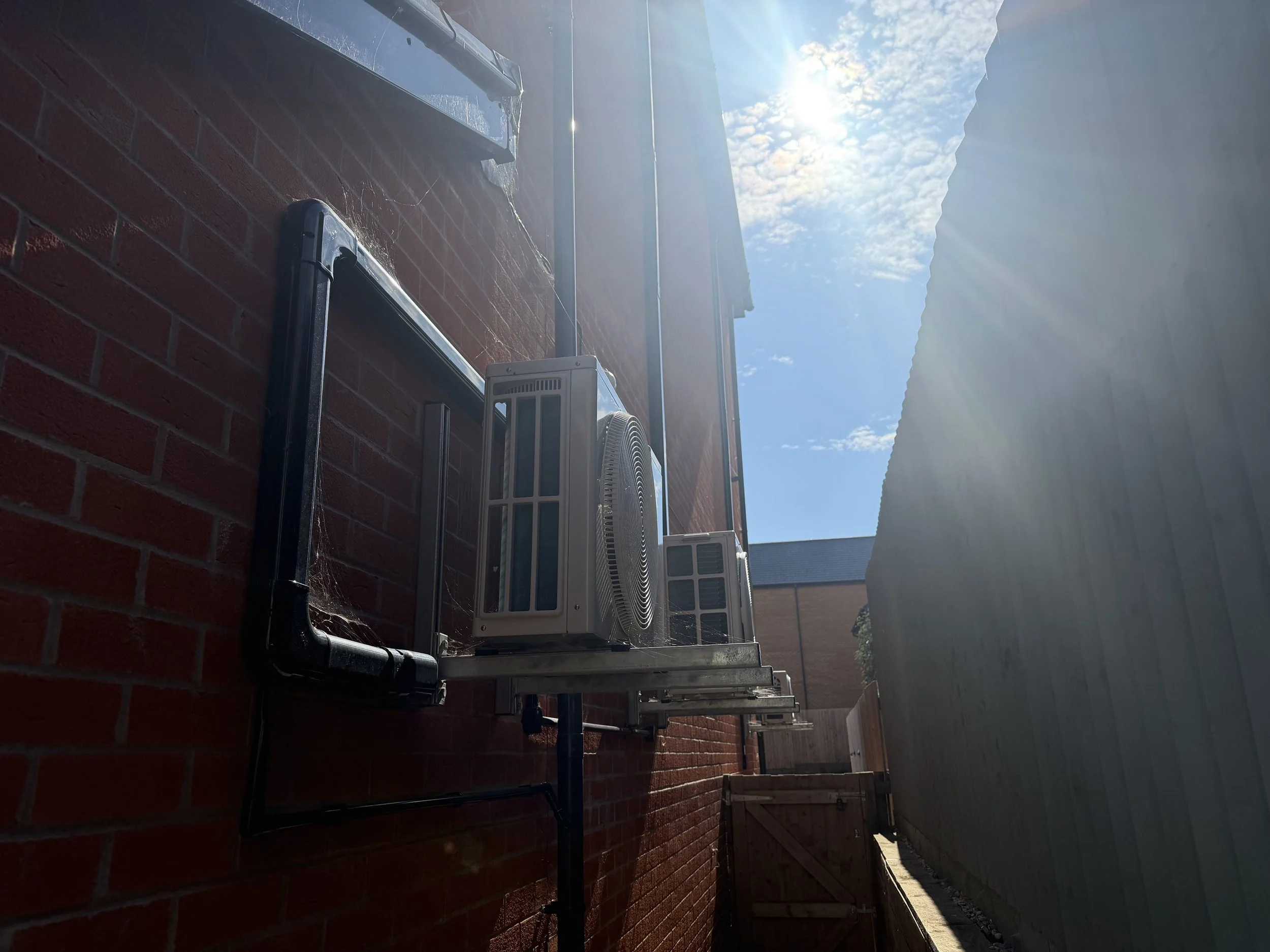 A row of air conditioning units installed on a brick wall outdoors, with a blue sky and sunlight shining down.