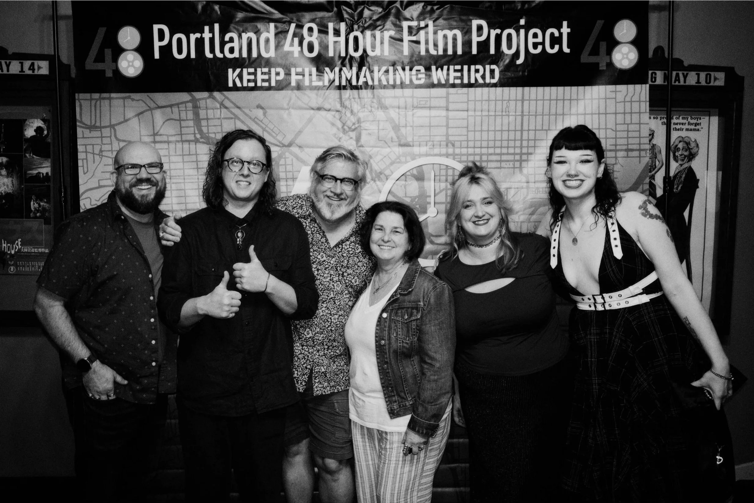 Group of seven people posing for a photo in front of a banner that reads 'Portland 48 Hour Film Project Keep Filmmaking Weird'. The group includes men and women of various ages, all smiling and standing close together.