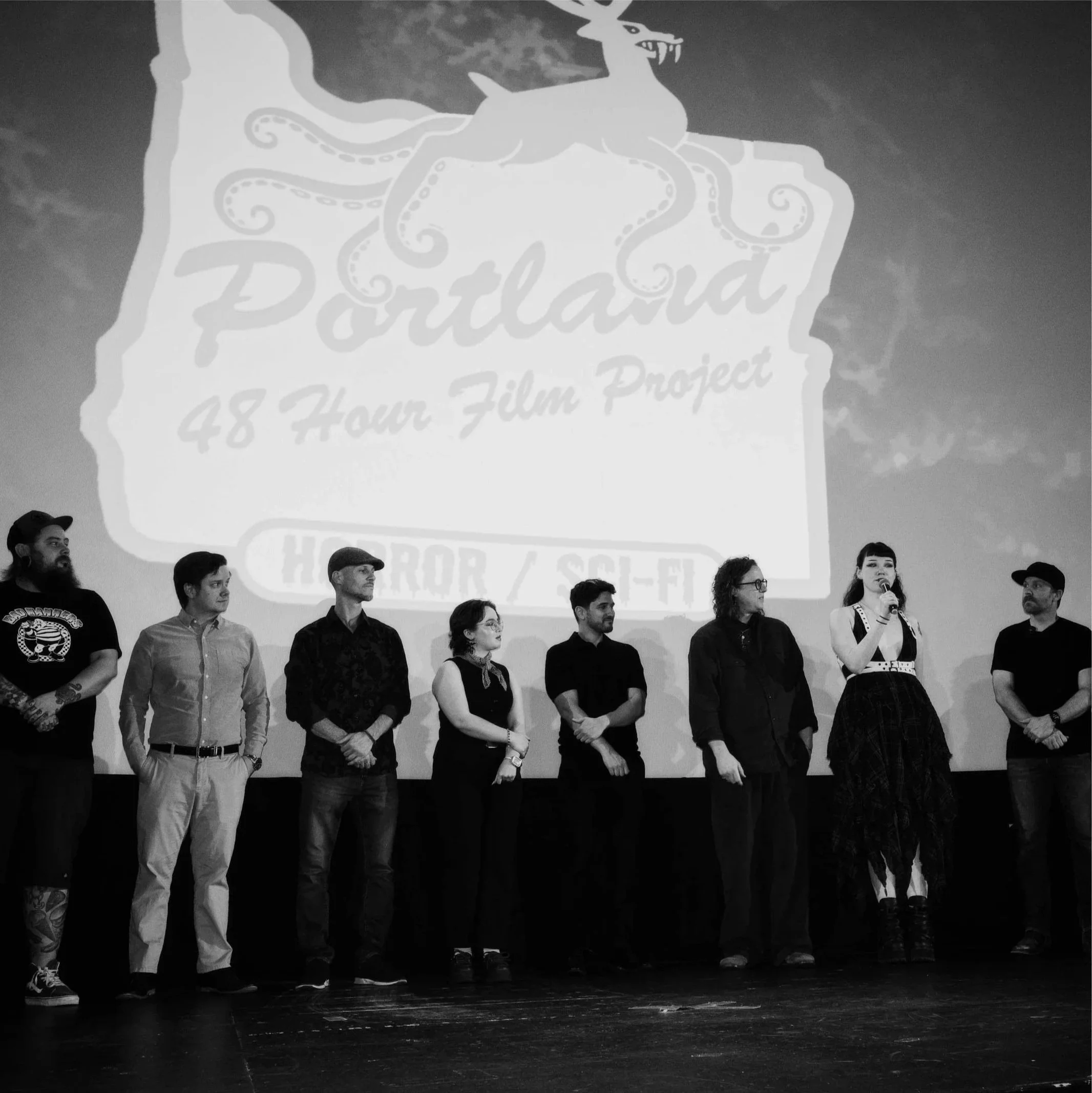 A group of people standing on stage with a large screen in the background displaying a logo that reads 'Portland 48 Hour Film Project' and an illustration of a dragon. The people appear to be participating in an event, possibly an award ceremony or presentation.