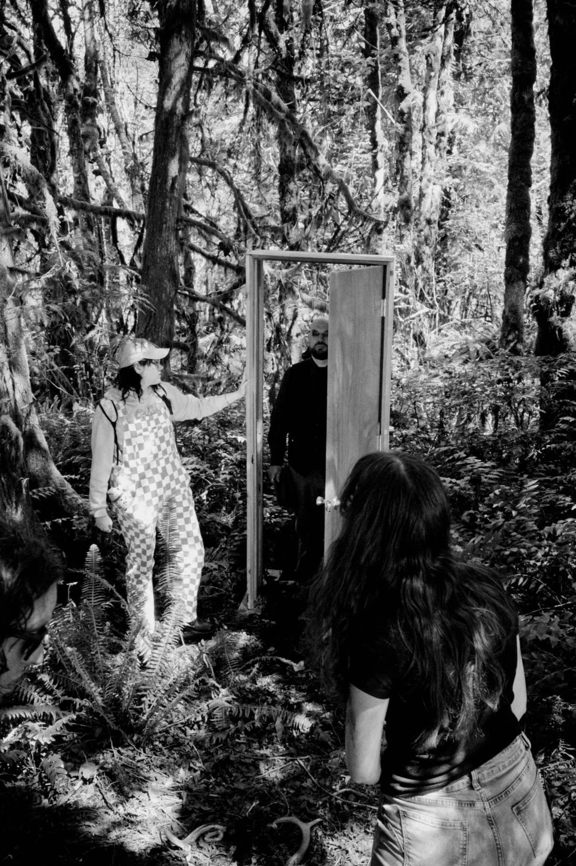 People in a forest, one woman with a checkered shirt and hat, another woman with long hair, and a man in black holding a door open.