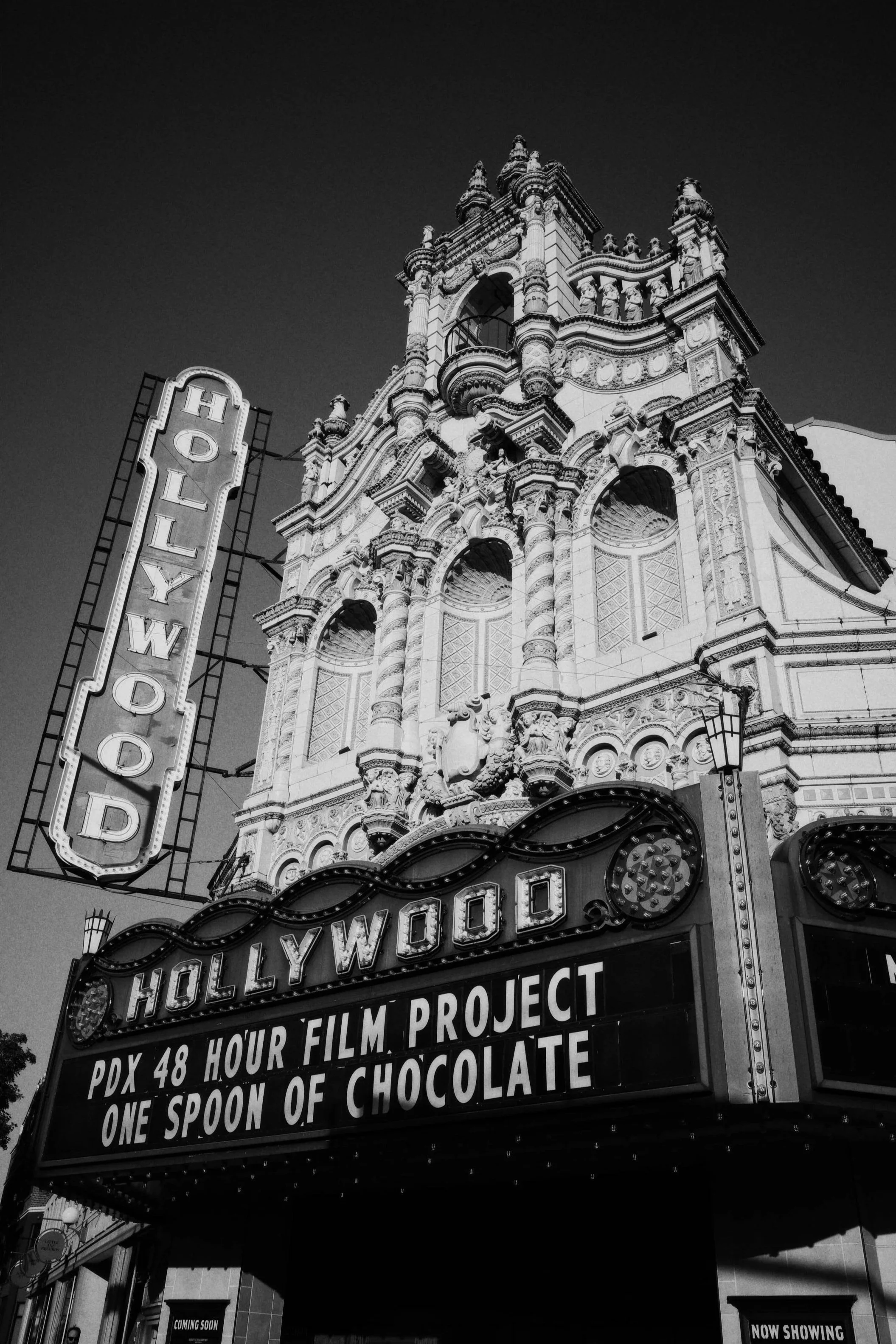 Black and white photo of a historic theater building with ornate architecture and a marquee sign advertising a film project, with a vertical lighted sign reading 'HOLLYWOOD' next to it.