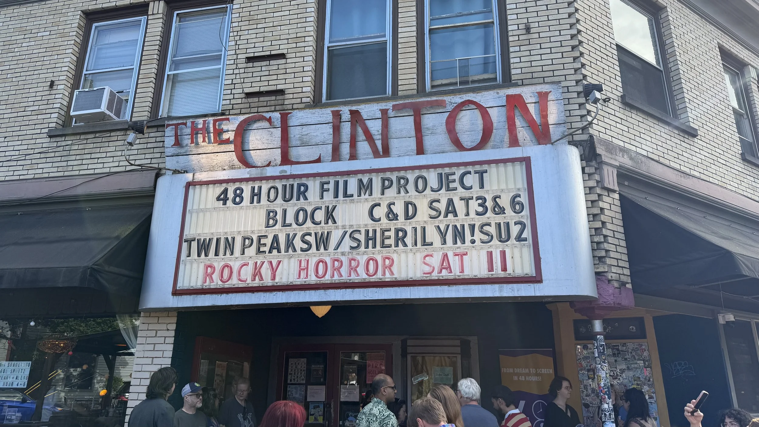 The marquee of the Clinton theater displays information about upcoming films and events, with a crowd of people gathered outside.