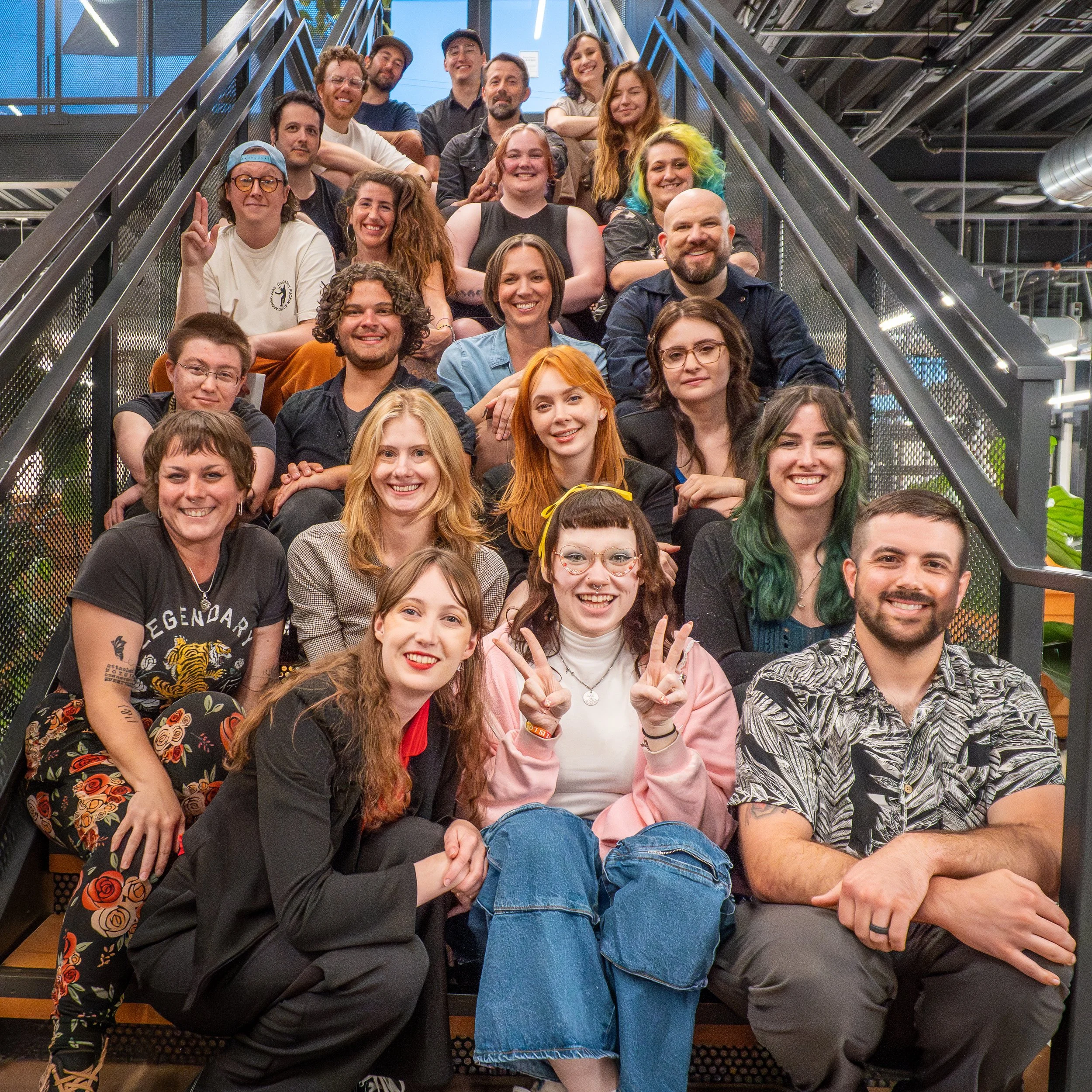 A diverse group of people, smiling and sitting on stairs made of metal, inside an industrial-style building with metal beams and stairs, in a casual group photo.