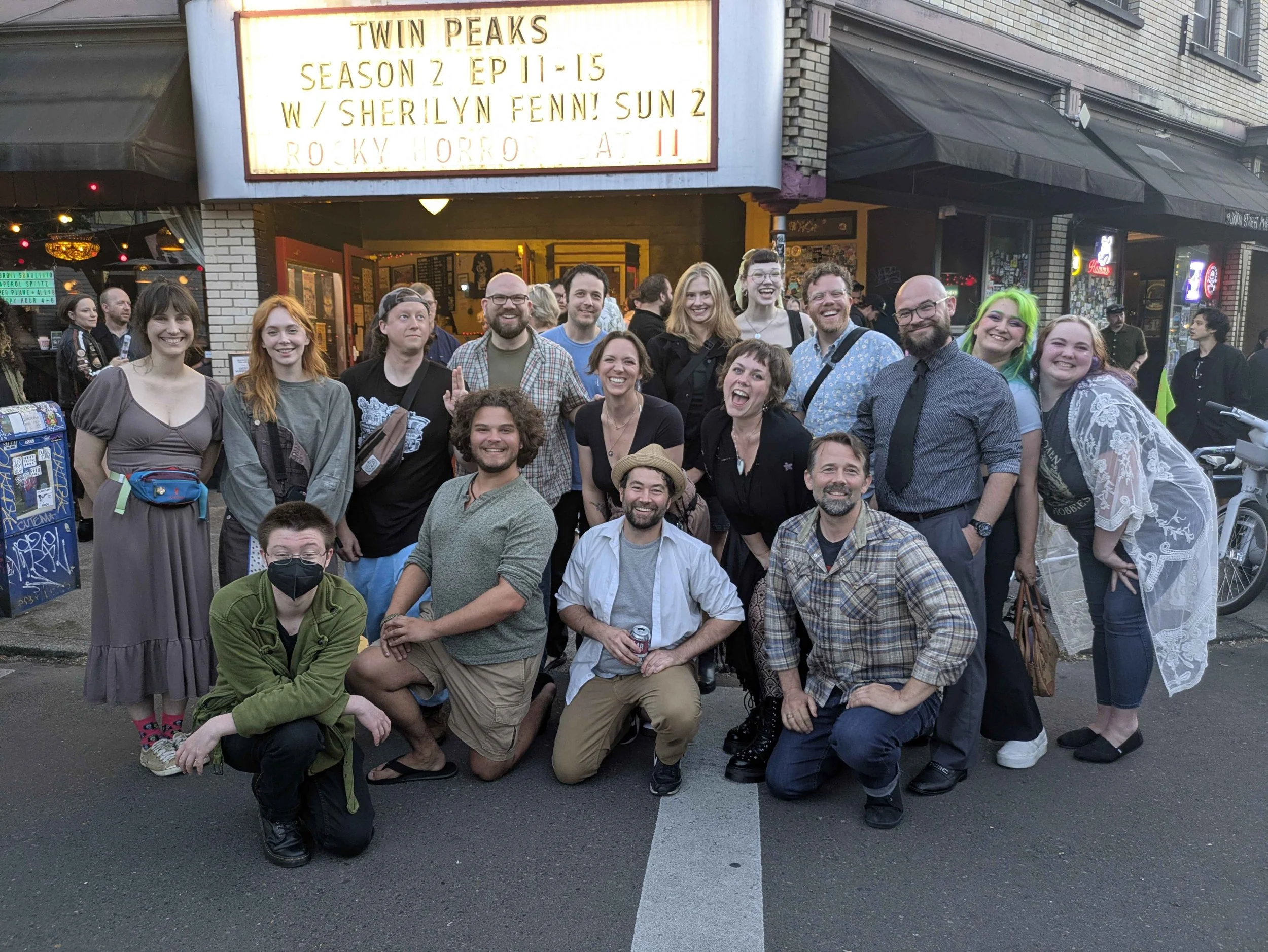 A group of people smiling, posing, and sitting in front of a comedy club with a marquee sign for Twin Peaks. The sign mentions season 2, episodes 11-15, with Sherilyn Fenn, on a city street at night.