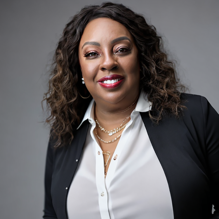 A woman with curly hair smiling at the camera, wearing a white blouse, black blazer, and layered necklaces, against a gray background.