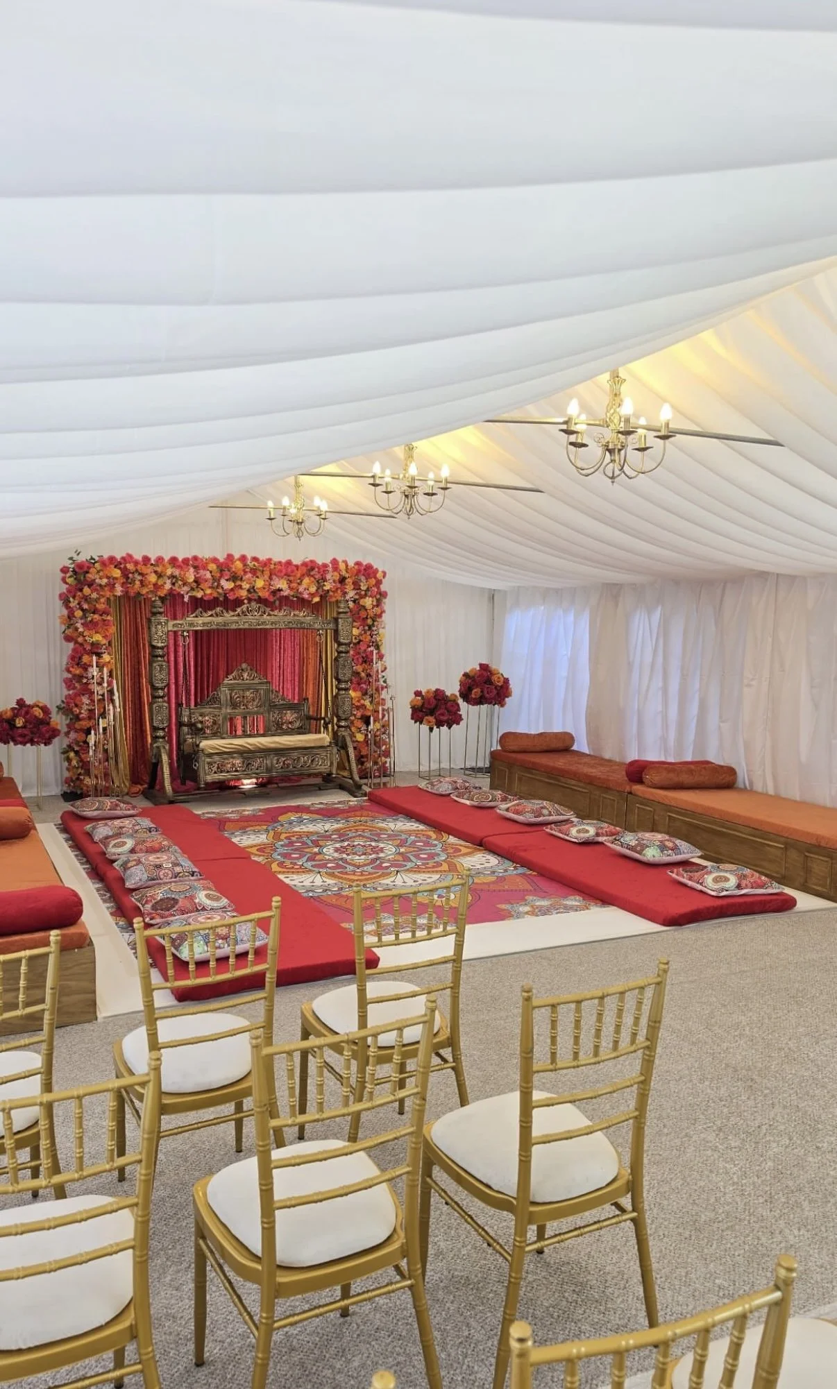 A decorated indoor wedding stage with a white fabric ceiling, chandeliers, a floral backdrop with pink and red flowers, a carved wooden throne, red cushions, and floral arrangements, surrounded by chairs with gold frames and white cushions.