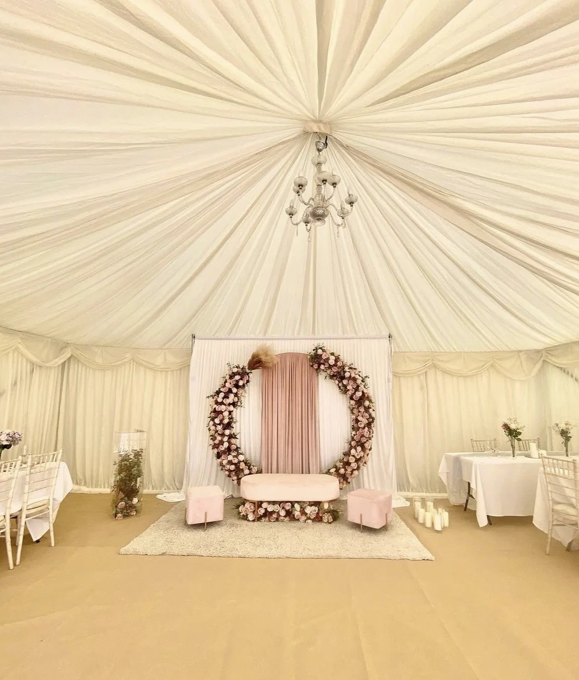 Elegant decorated event tent with a chandelier, floral arch, pink and white seating, and tables with flowers.
