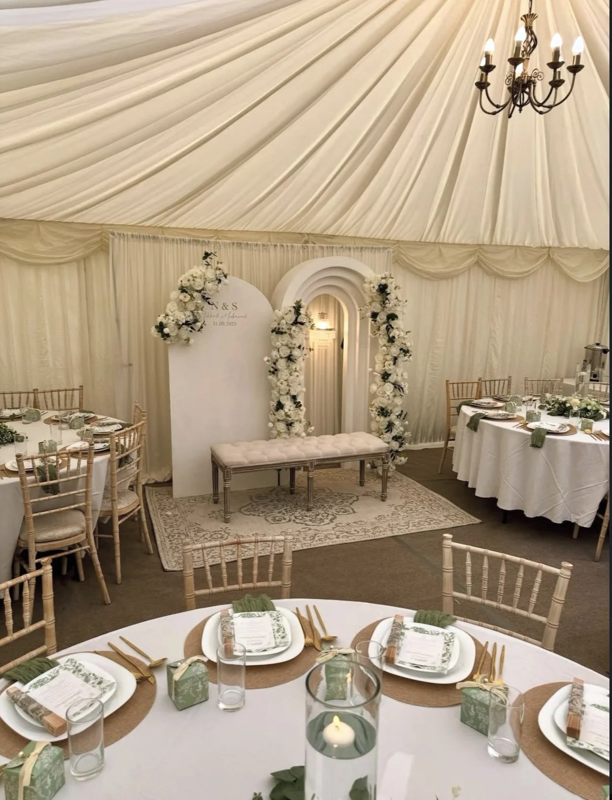 Elegant wedding reception inside a cream-colored tent with draped fabric ceiling, decorated with a chandelier and floral arch, featuring round tables with white tablecloths, gold chairs, and place settings.