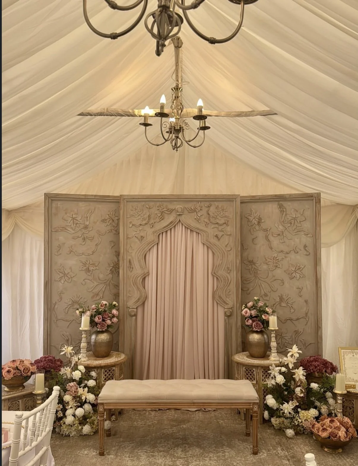 Elegant wedding altar with a fabric-draped ceiling and a chandelier. The backdrop features detailed carved wood with floral and vine patterns, flanked by vases of flowers and candles on side tables, and a bench in front.
