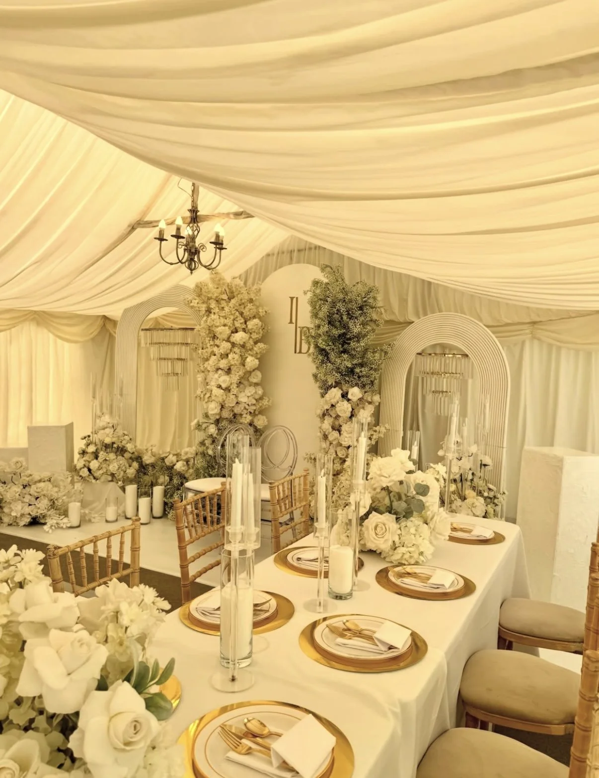 Elegant wedding reception setup with white floral arrangements, tall candles, and gold table settings inside a tent with cream drapery.