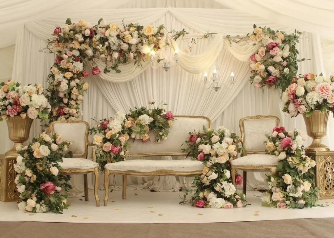 Elegant wedding ceremony backdrop with floral arrangements of pink, peach, and white roses, accompanied by cream-colored drapes and gold furniture.