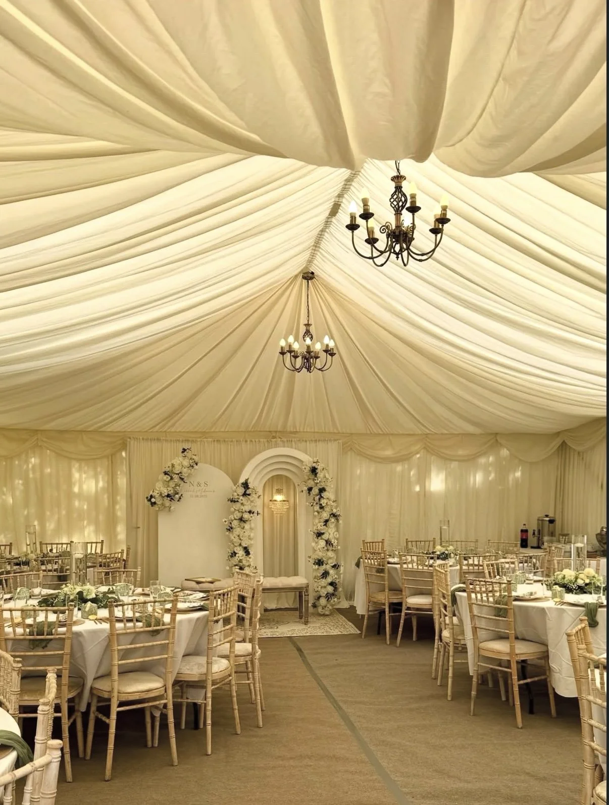 Elegant wedding reception inside a decorated tent with a draped ceiling, chandeliers, round tables with white tablecloths, and a floral archway.