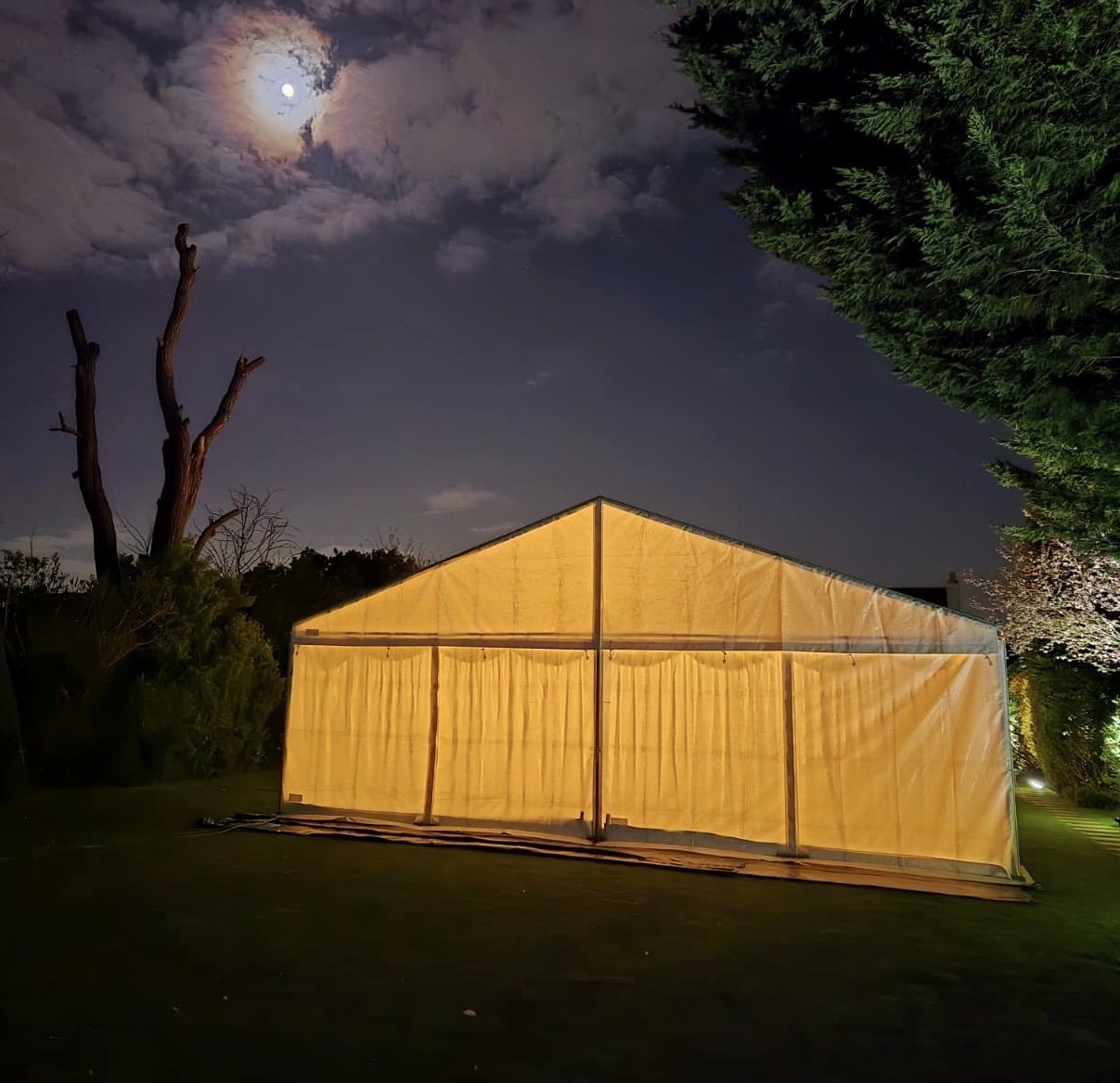 A large yellow tent set up outdoors at night, illuminated from within, with a full moon and clouds in the sky, surrounded by trees and foliage.