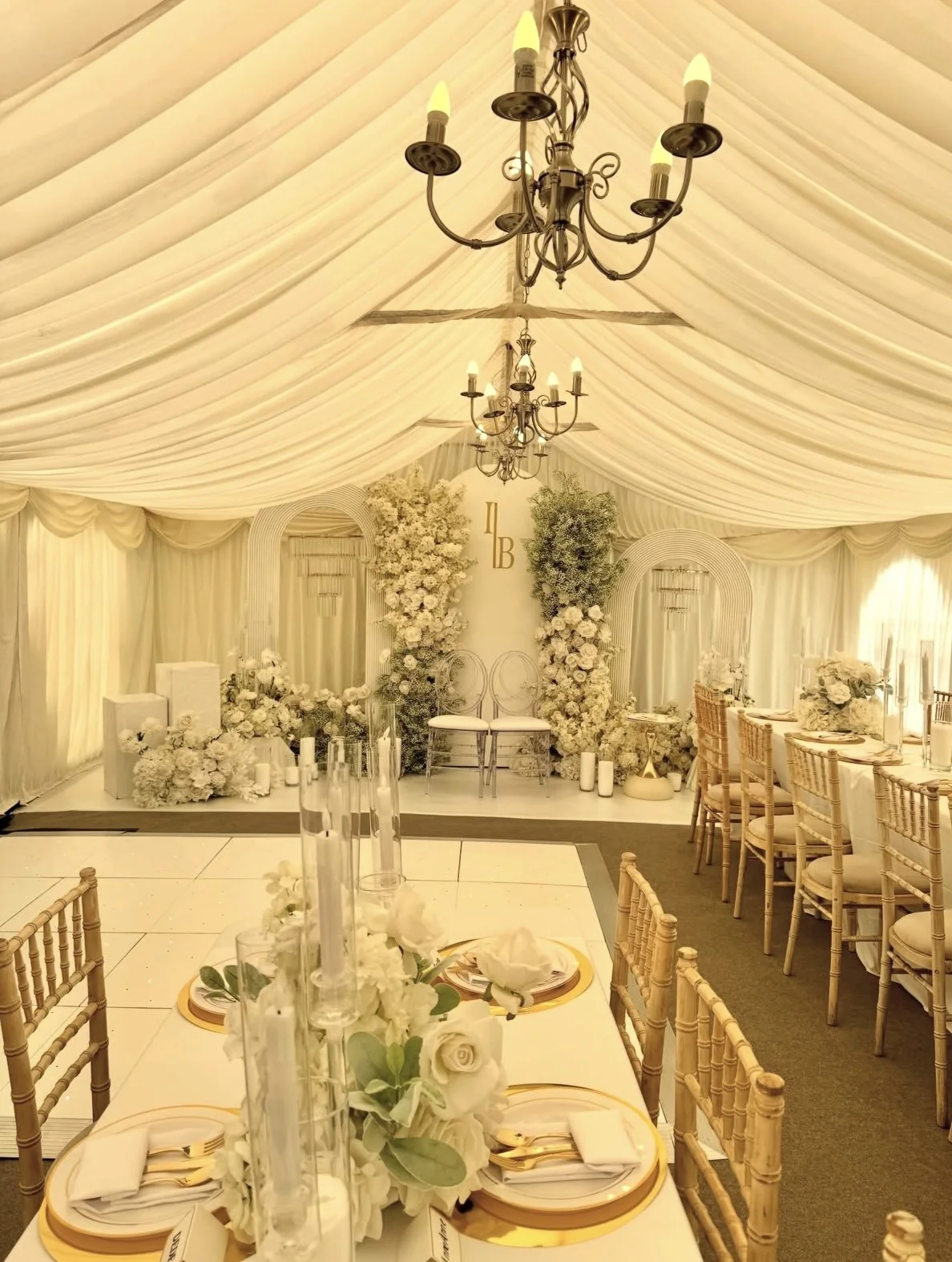 Elegant wedding reception inside a cream-colored tent with draped fabric and chandeliers. The decor features white and light green floral arrangements, candles, and a seating area with two chairs in front of a monogrammed backdrop with the initials '