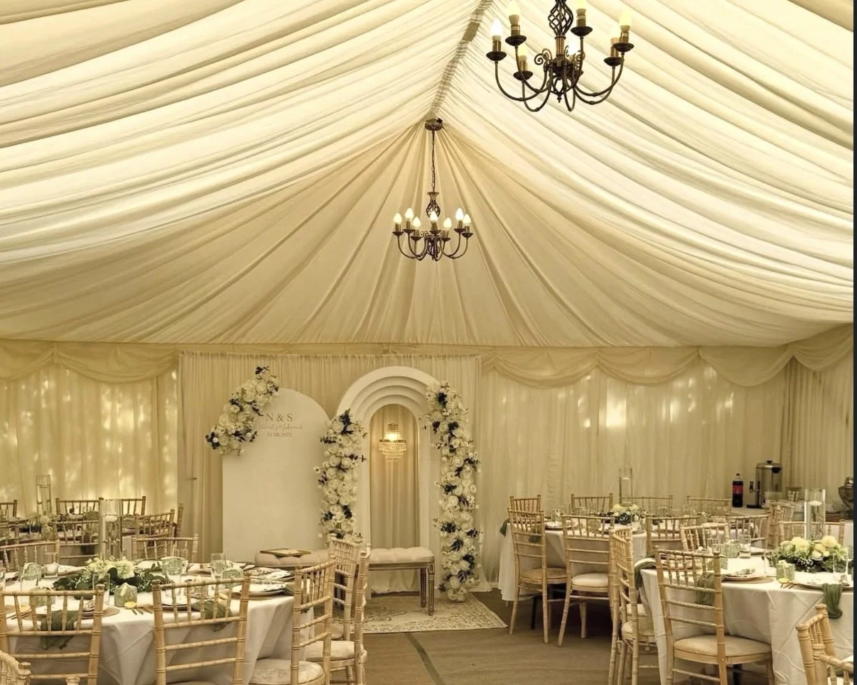 Elegant wedding reception inside a large cream-colored tent with draped fabric ceiling, chandeliers, round tables with white tablecloths and floral centerpieces, and an arch decorated with white flowers.