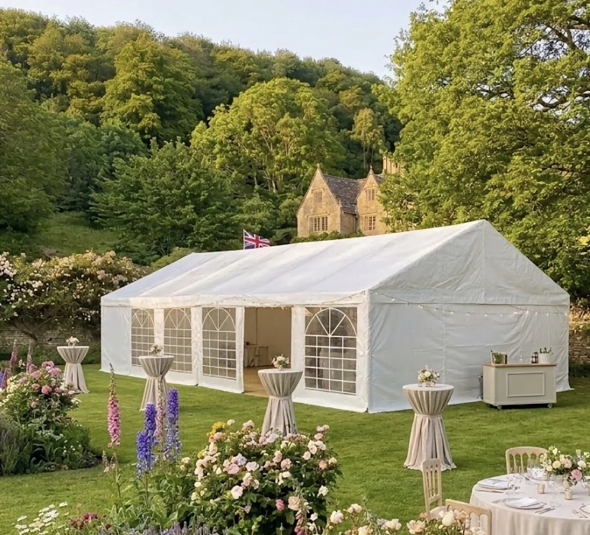 A large white event tent set up on a grassy lawn, decorated with flowers and surrounded by tables and chairs for a celebration, with green trees and a stone building with a Union Jack flag in the background.