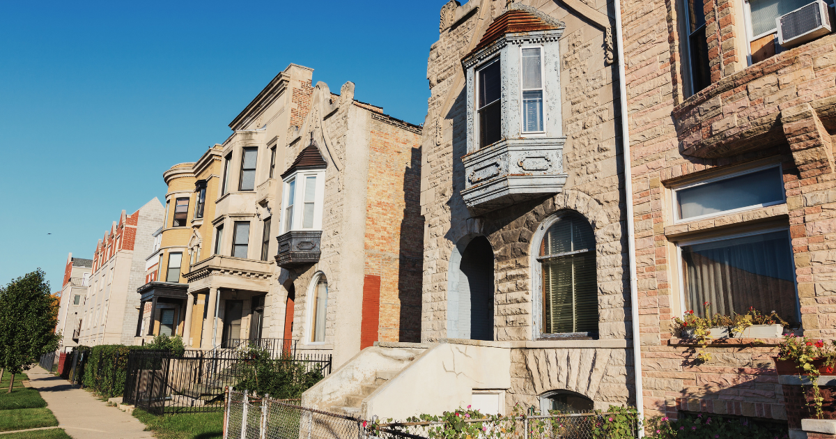 Row of historic residential buildings with brick and stone facades, bay windows, and small front yards along a sidewalk under a clear blue sky.