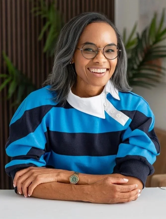 A woman with shoulder-length gray hair, glasses, and a bright smile, sitting at a table with her arms crossed, wearing a blue and black striped rugby shirt and a watch, with a background of plants.