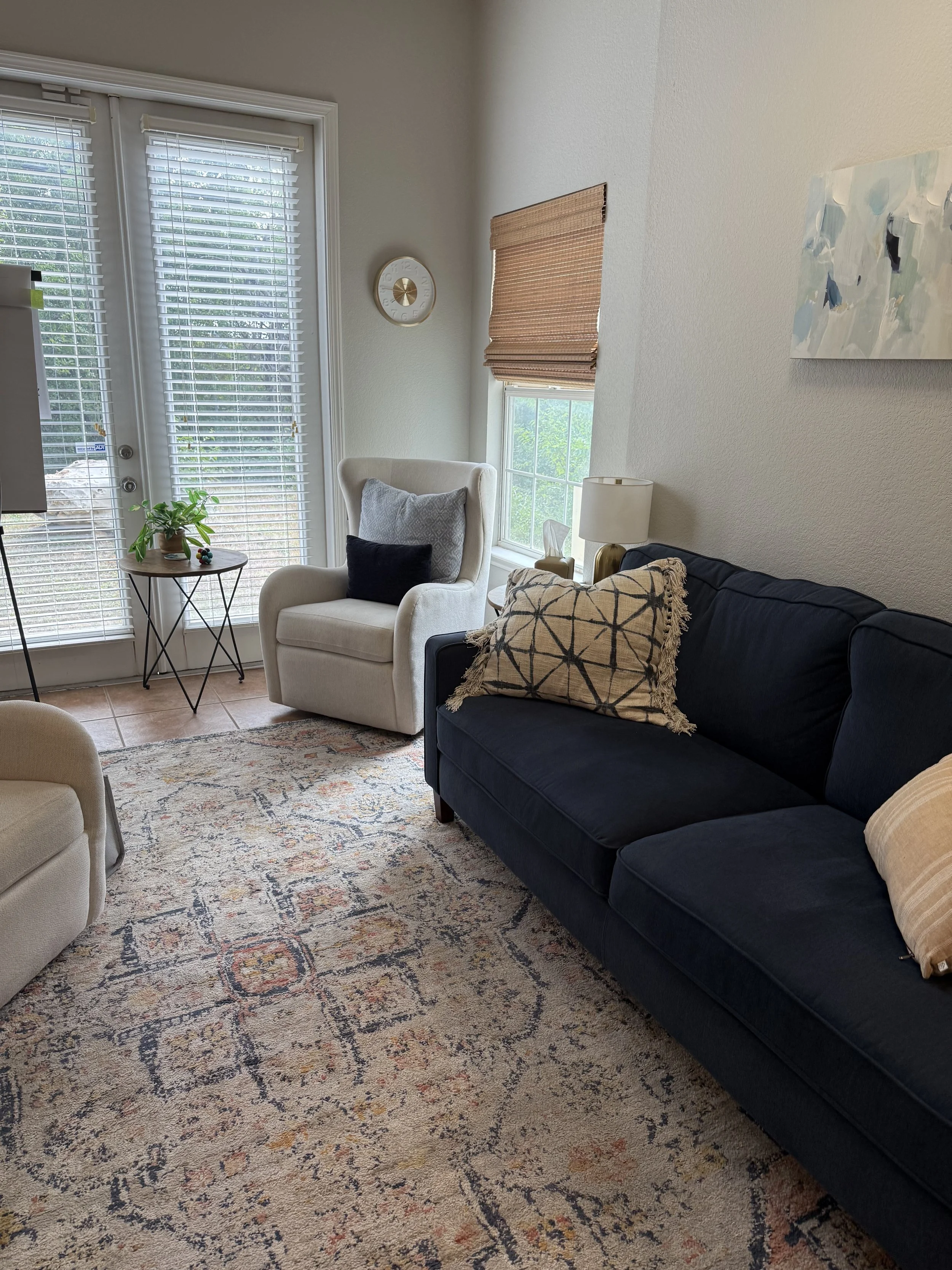 A cozy living room featuring a navy blue couch dressed with decorative pillows, a white armchair with a dark pillow, a patterned area rug, a small side table with a plant, a lamp, a wall clock, a window with a bamboo shade, and a sliding glass door with white blinds.