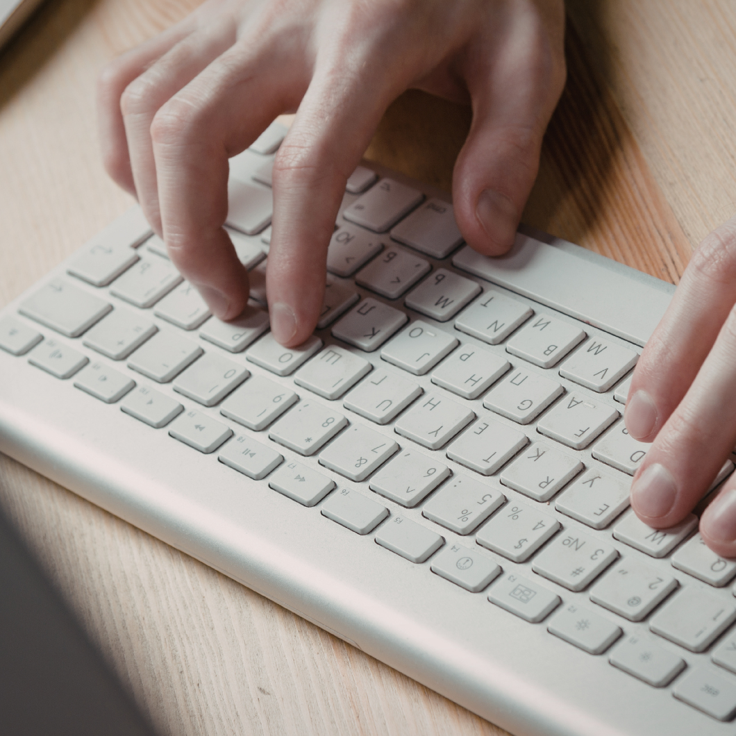 Close-up of a person typing on a white computer keyboard on a wooden desk.