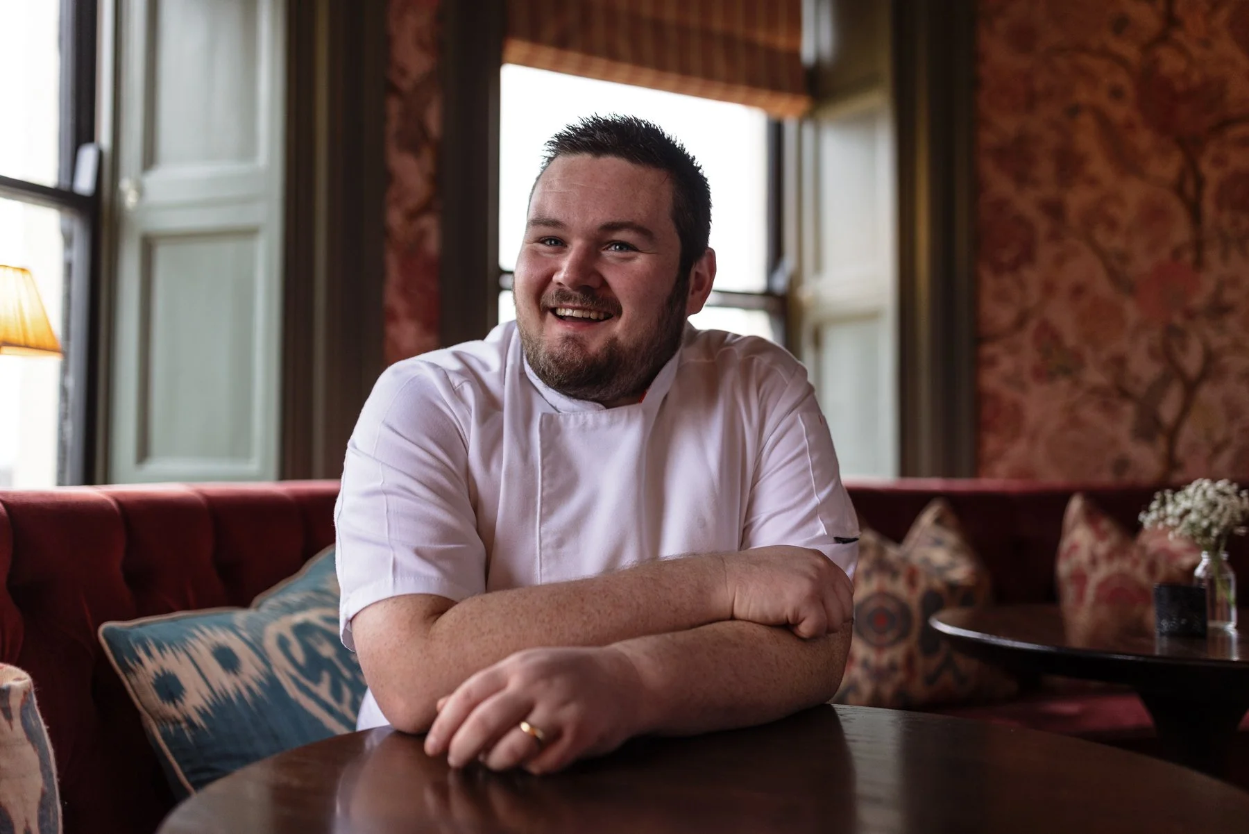 A smiling man wearing a white chef's coat sitting at a round wooden table in a cozy, vintage-style restaurant with plaid curtains and floral wallpaper.