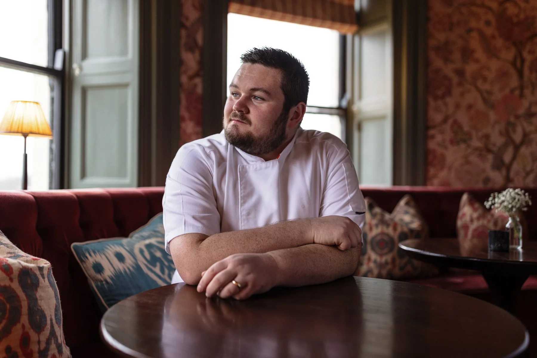 A man sits at a wooden table in a cozy, vintage-style restaurant or cafe, wearing a white chef's jacket, with his arms crossed on the table, looking off to the side.