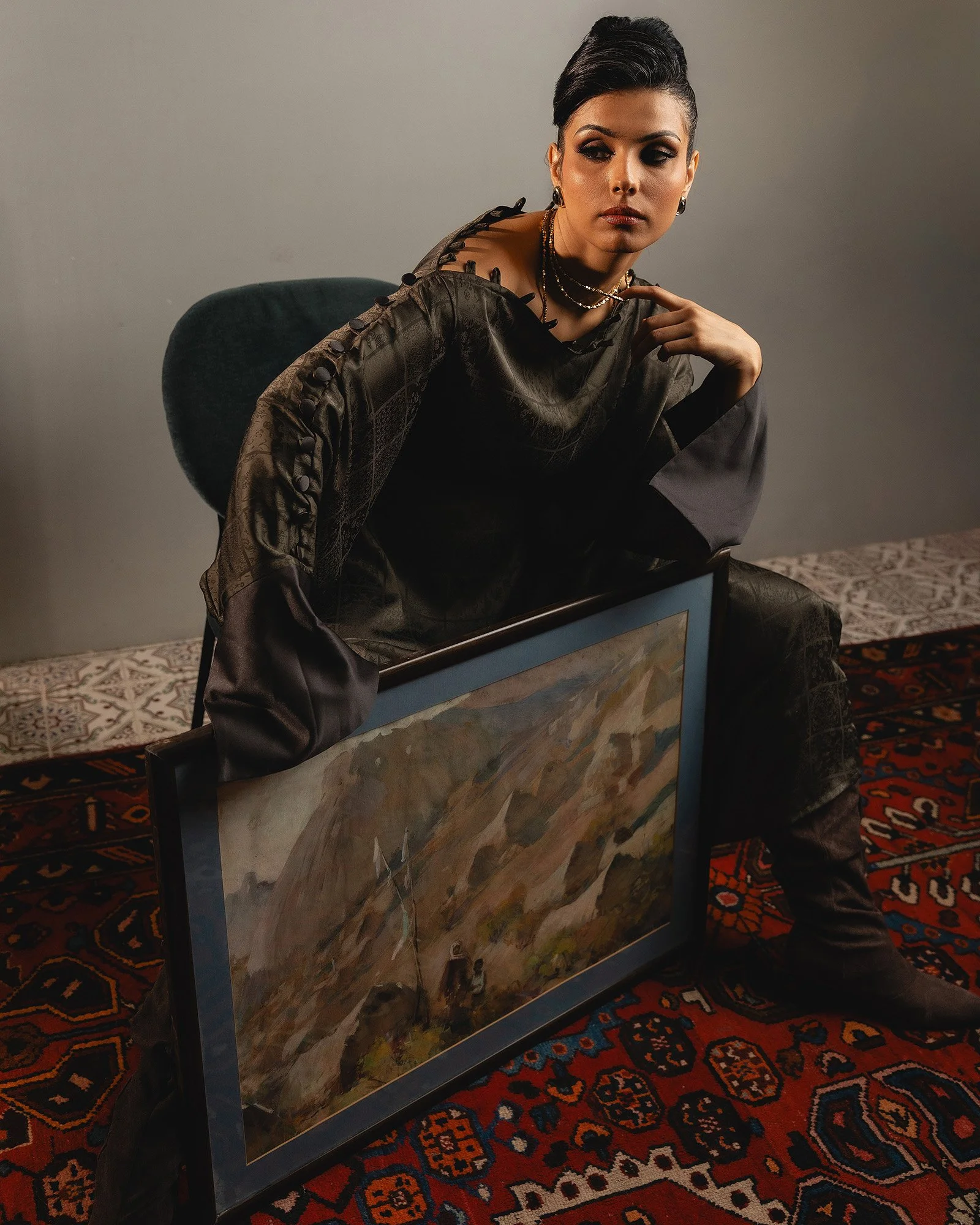 A woman sitting on a chair with a painting in front of her, covering her lap, in a room with patterned carpet. The woman has dark hair, wears dark makeup, and is dressed in dark, textured clothing with buttons and jewelry.
