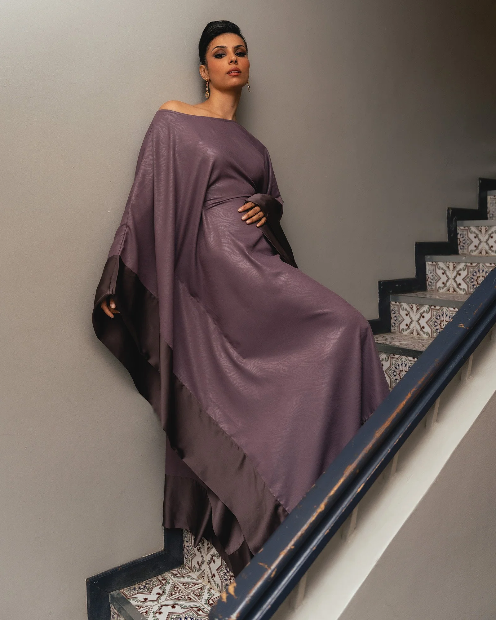A woman in a purple dress resting against a plain wall next to a staircase with patterned tiles.