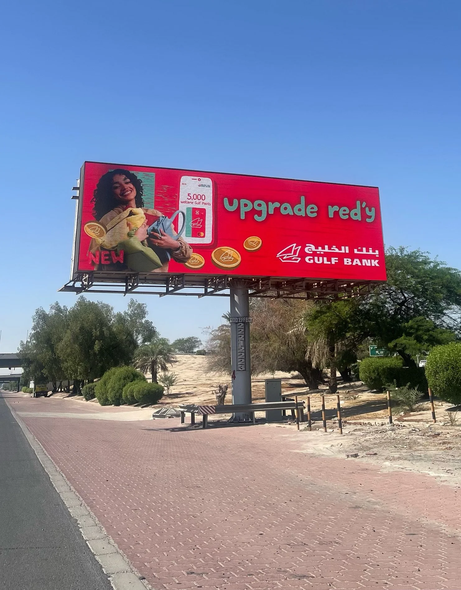 A billboard advertisement for Gulf Bank with a smiling woman holding a phone, displaying a message of 5,000 Gulf Points. The billboard has a red background with the Gulf Bank logo and the slogan 'Upgrade ready' in green letters.